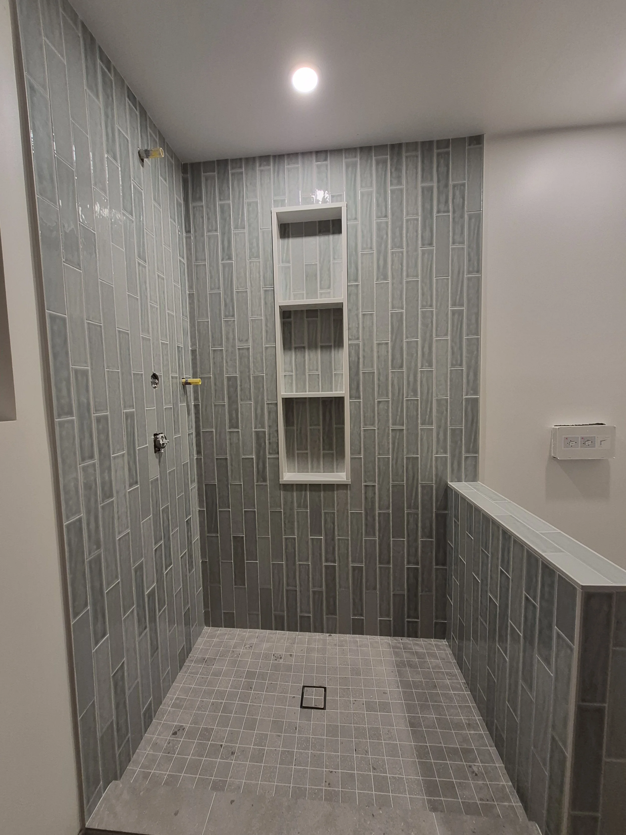 Unfinished bathroom shower with gray tile walls, small gray tile floor, built-in shelves, and plumbing fixtures not yet installed.