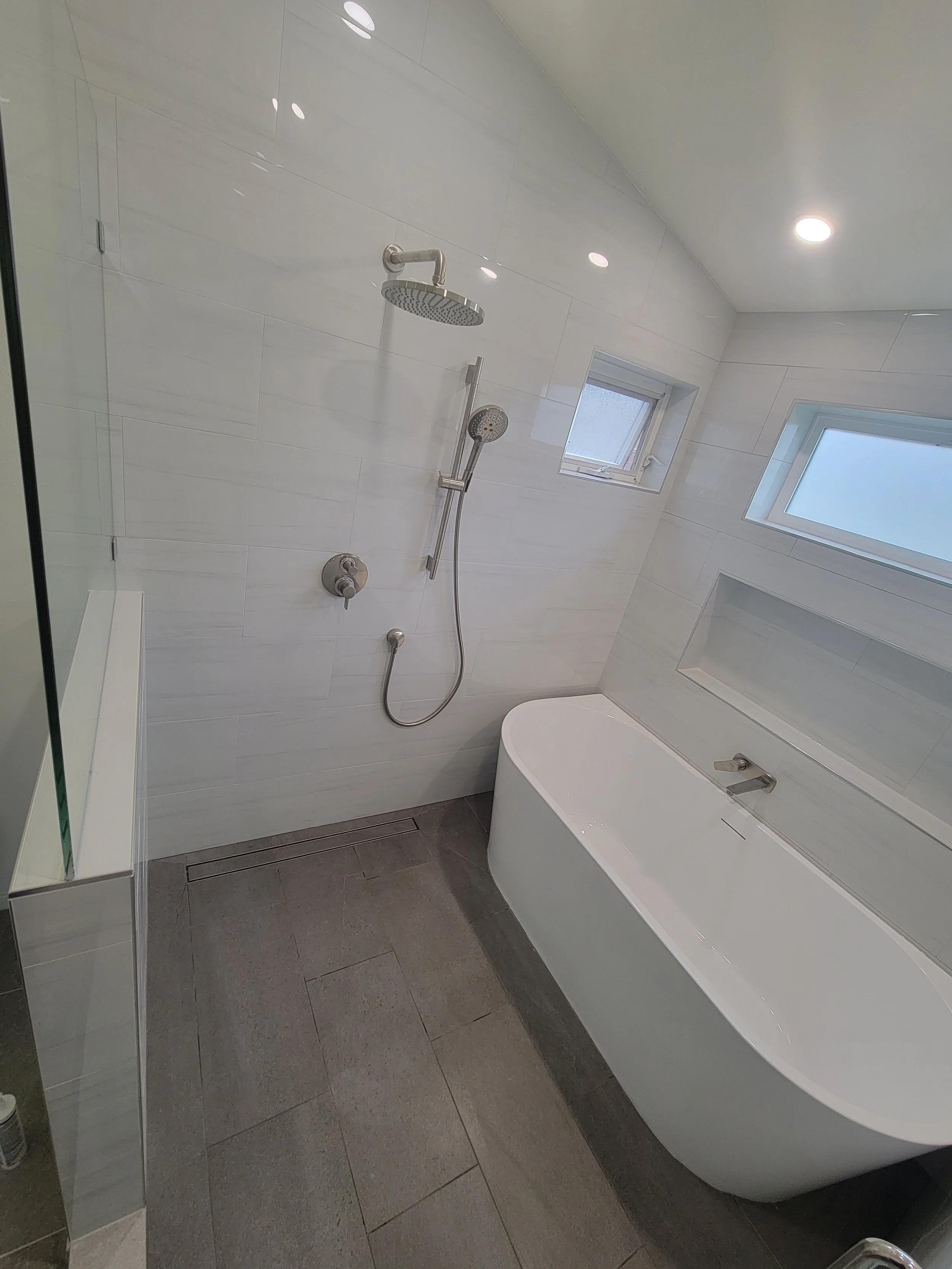 Modern bathroom with white tiled walls, a freestanding bathtub, two small high windows, and a wall shower with a rain showerhead and handheld shower attachment.