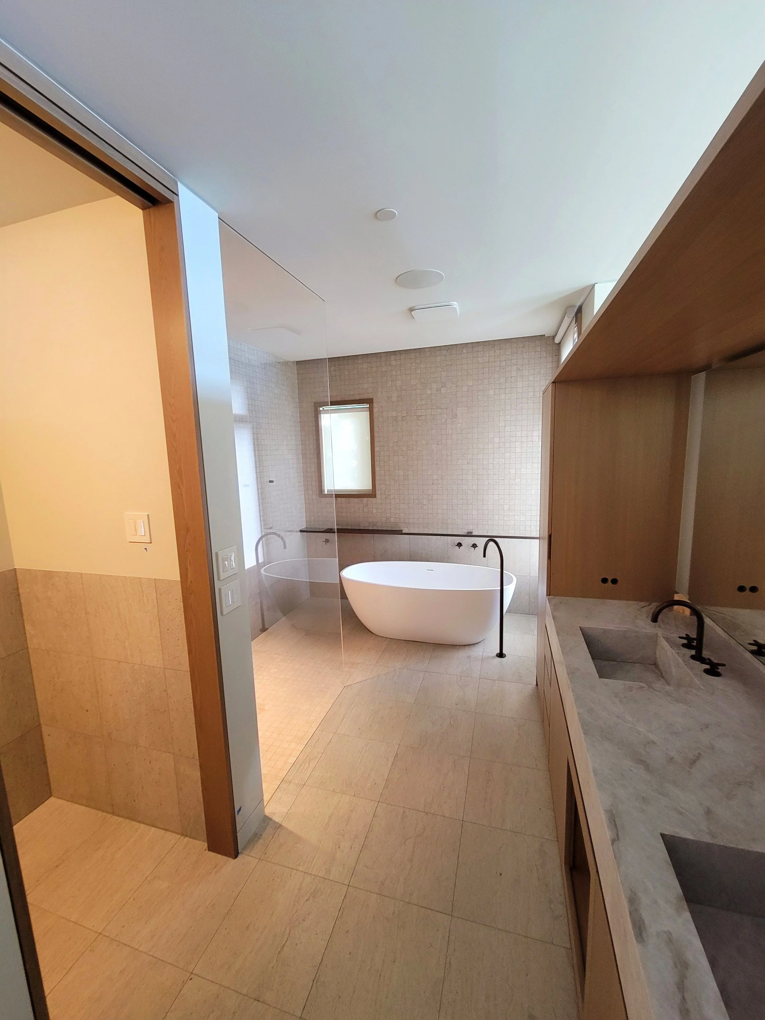 Modern bathroom with a freestanding bathtub near a window, tiled walls, and a marble countertop with sinks.