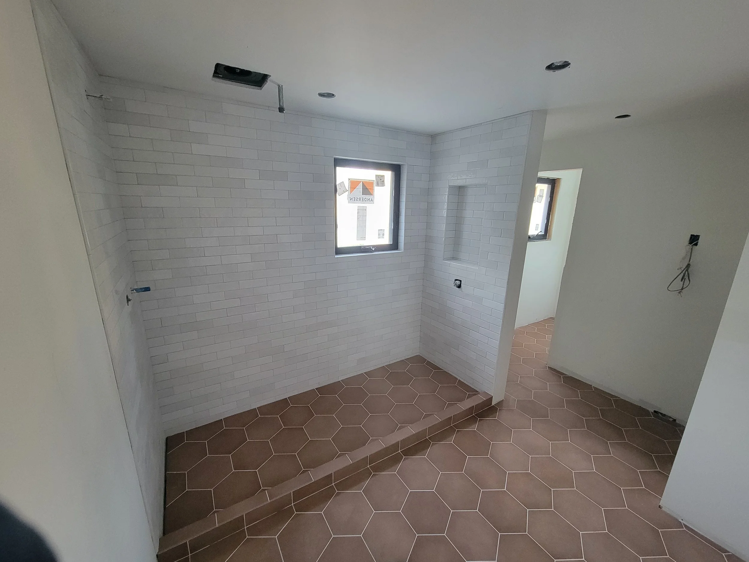 Empty room with hexagon brown floor tiles, white brick walls, and small windows with construction signs. There are unfinished ceiling fixtures and exposed wiring.