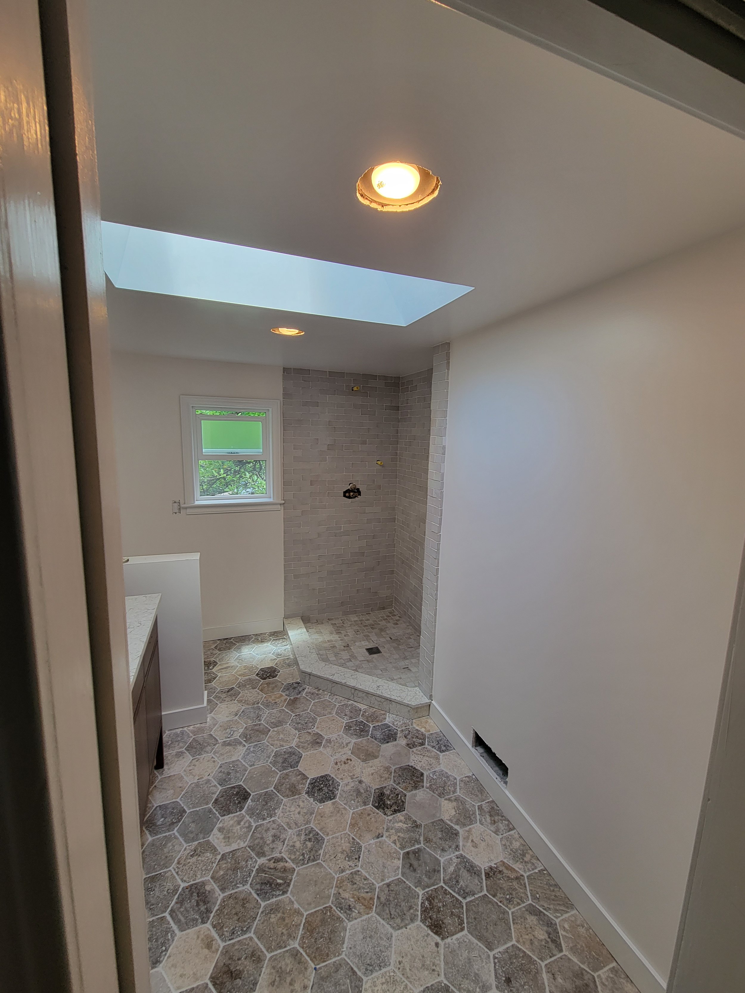 Bathroom under renovation showing hexagonal tile flooring, a small window, a skylight, and an empty walk-in shower area with gray tiles on the wall.
