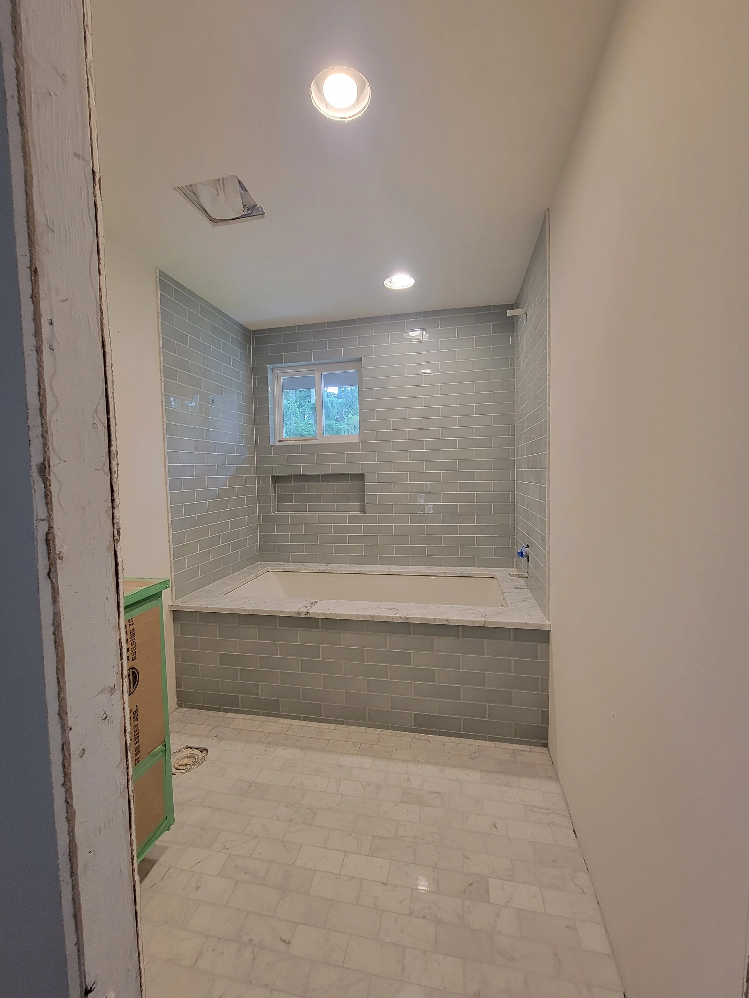 Bathroom under construction with gray tiled walls, a built-in bathtub, small window, and recessed ceiling lights.