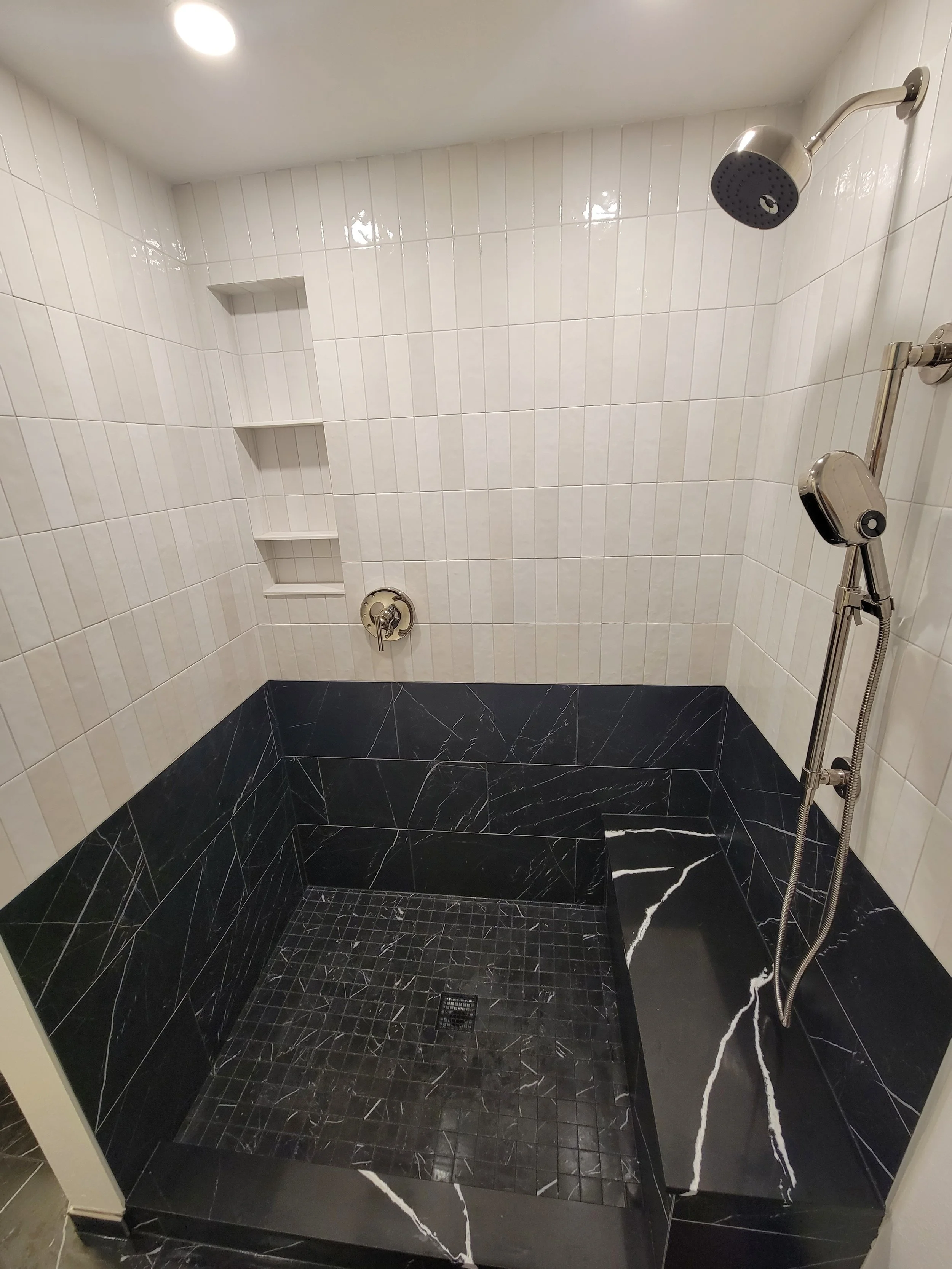 Modern shower area with white tiled walls, black marble tiles on the floor and lower walls, a rain showerhead, a handheld showerhead, built-in wall shelves, and a drain on the tiled floor.