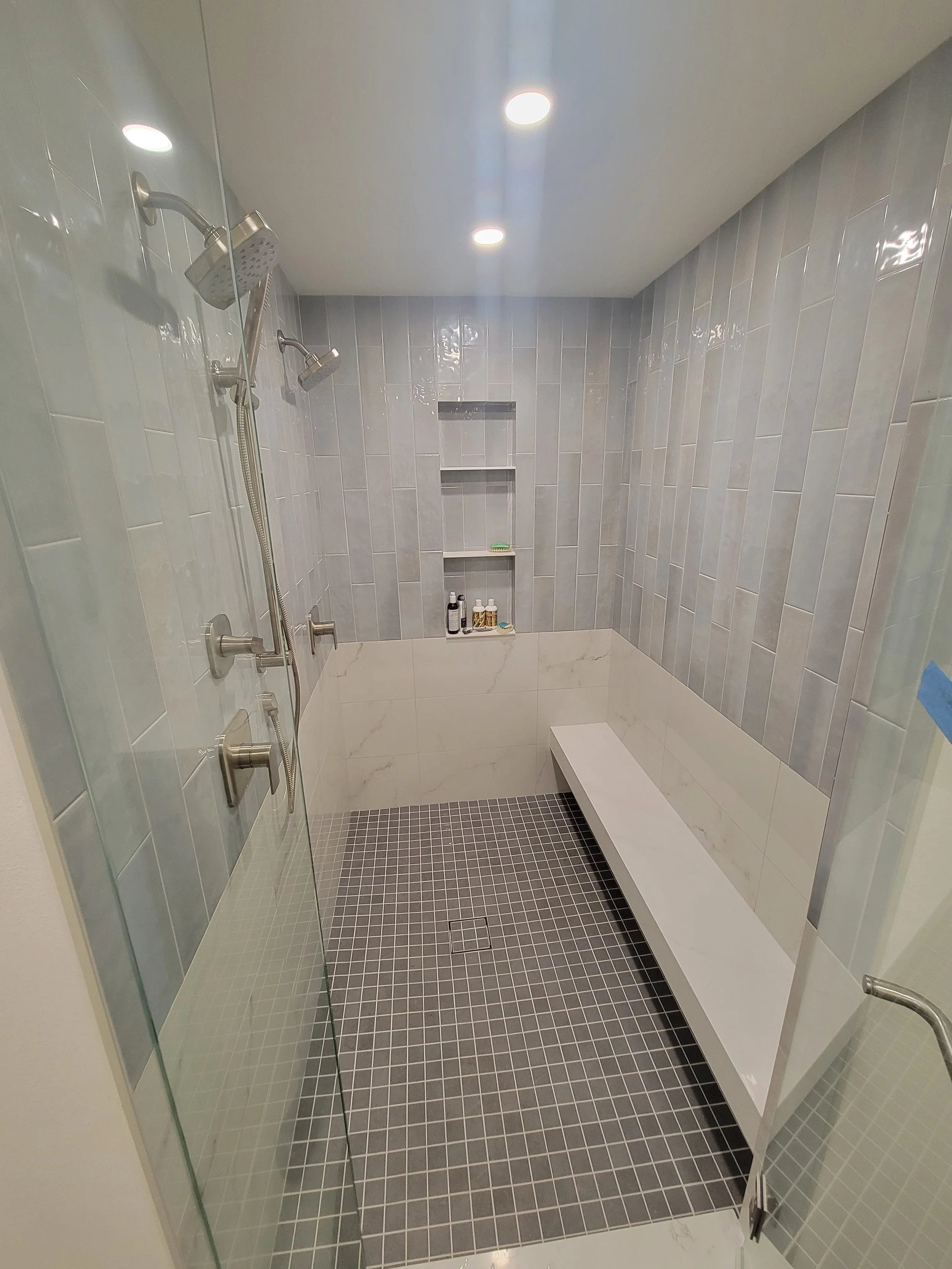 Modern shower with gray tiles on walls, black square tiles on floor, glass door, built-in bench, and built-in shelves with toiletries.