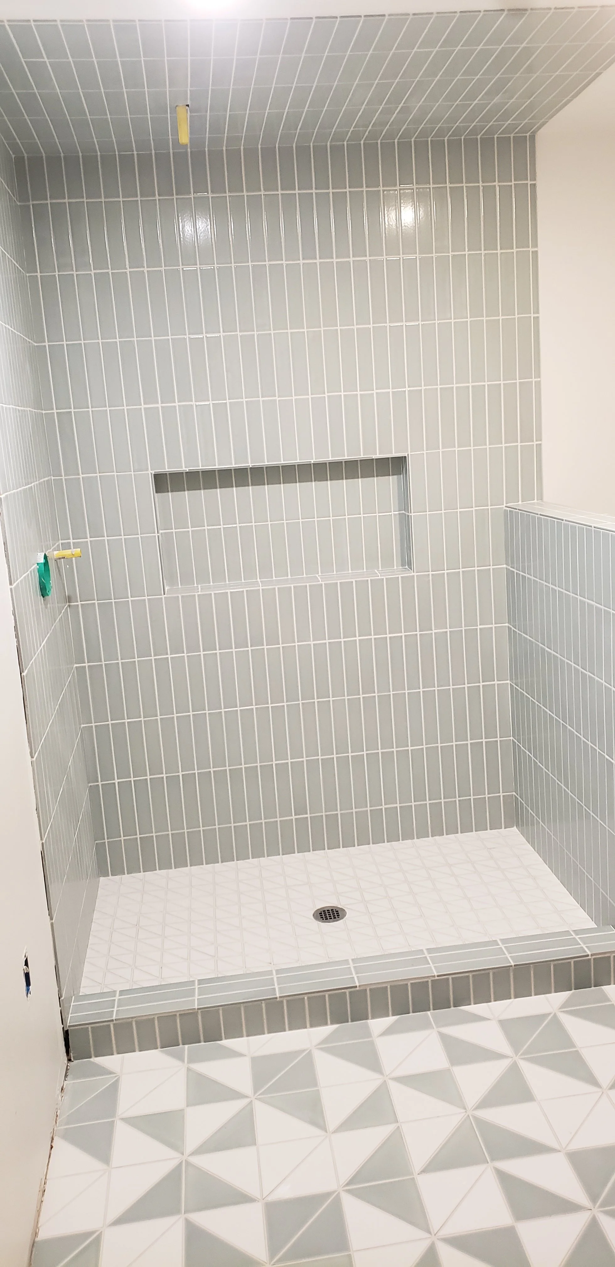 Empty shower stall with gray tiled walls, built-in niche, and a drain in the tiled floor. Part of the bathroom floor with gray and white geometric tiles is visible in the foreground.