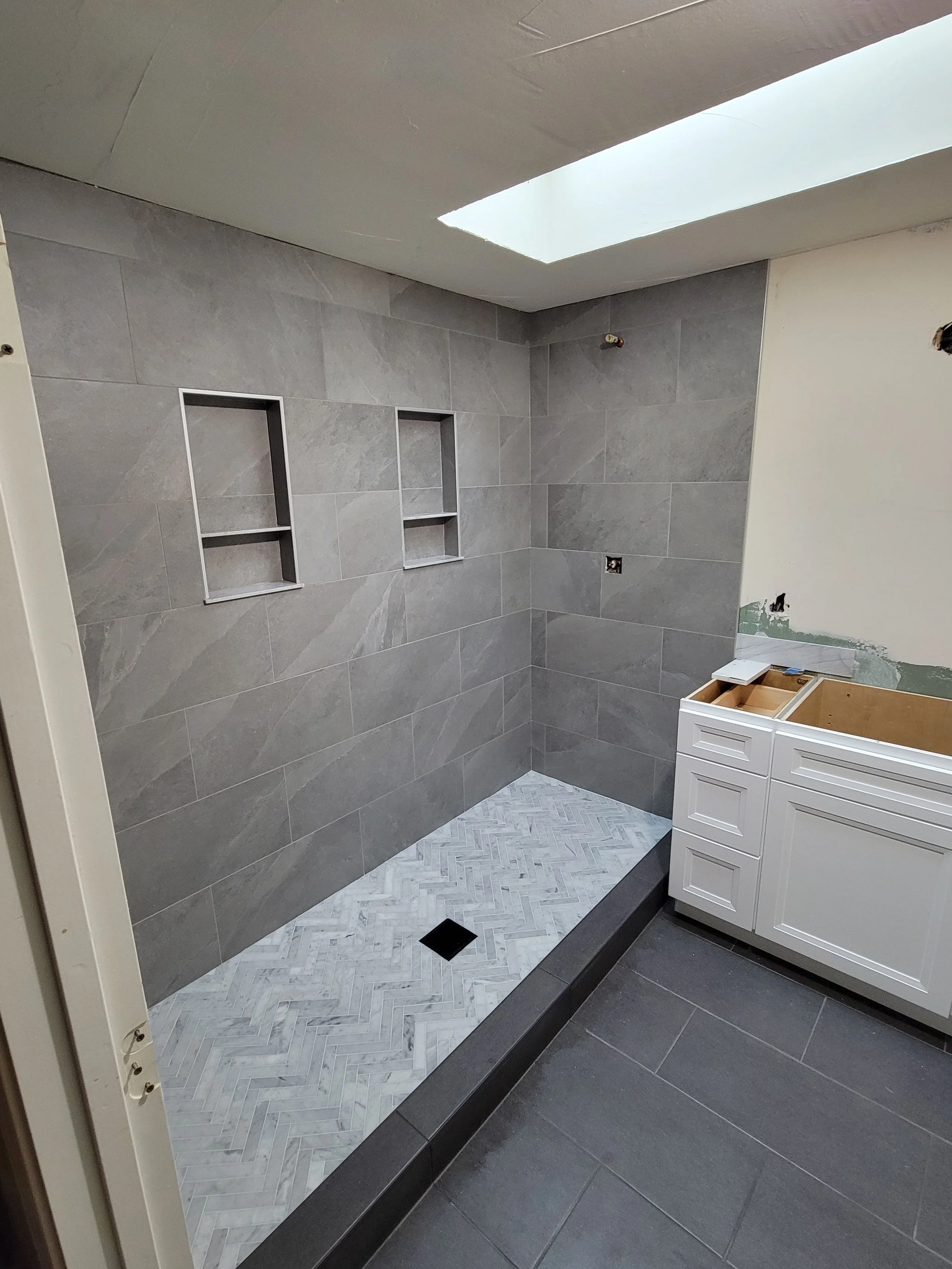 Bathroom still under construction with gray tiled walls, two recessed shelves in the wall, a skylight, and a partially installed white vanity cabinet.