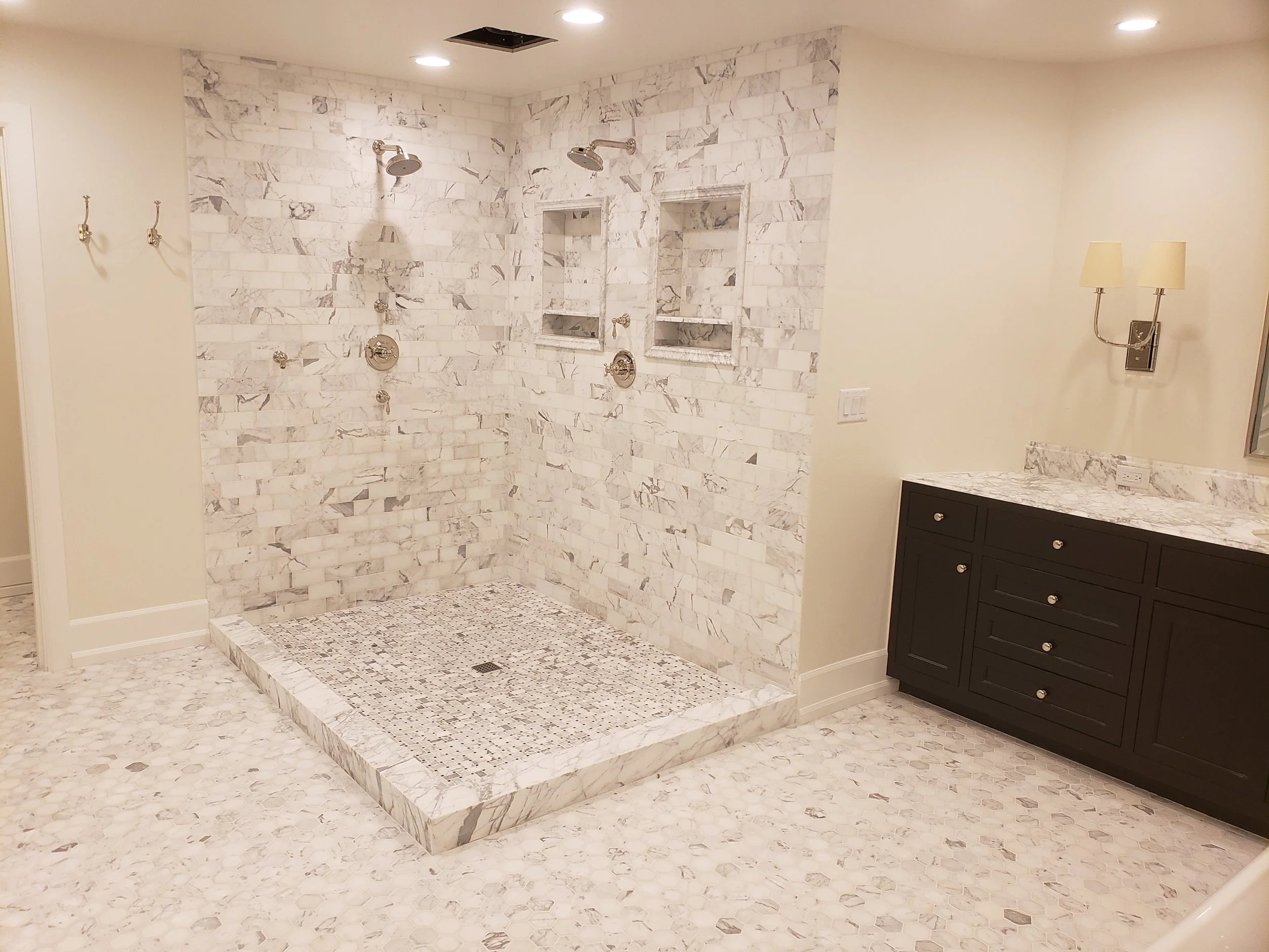 A spacious bathroom with a large walk-in shower featuring white marble tiles, built-in niches, dual shower heads, and a tiled shower base. There is a dark wood vanity with a marble countertop and a double sconce light above it.