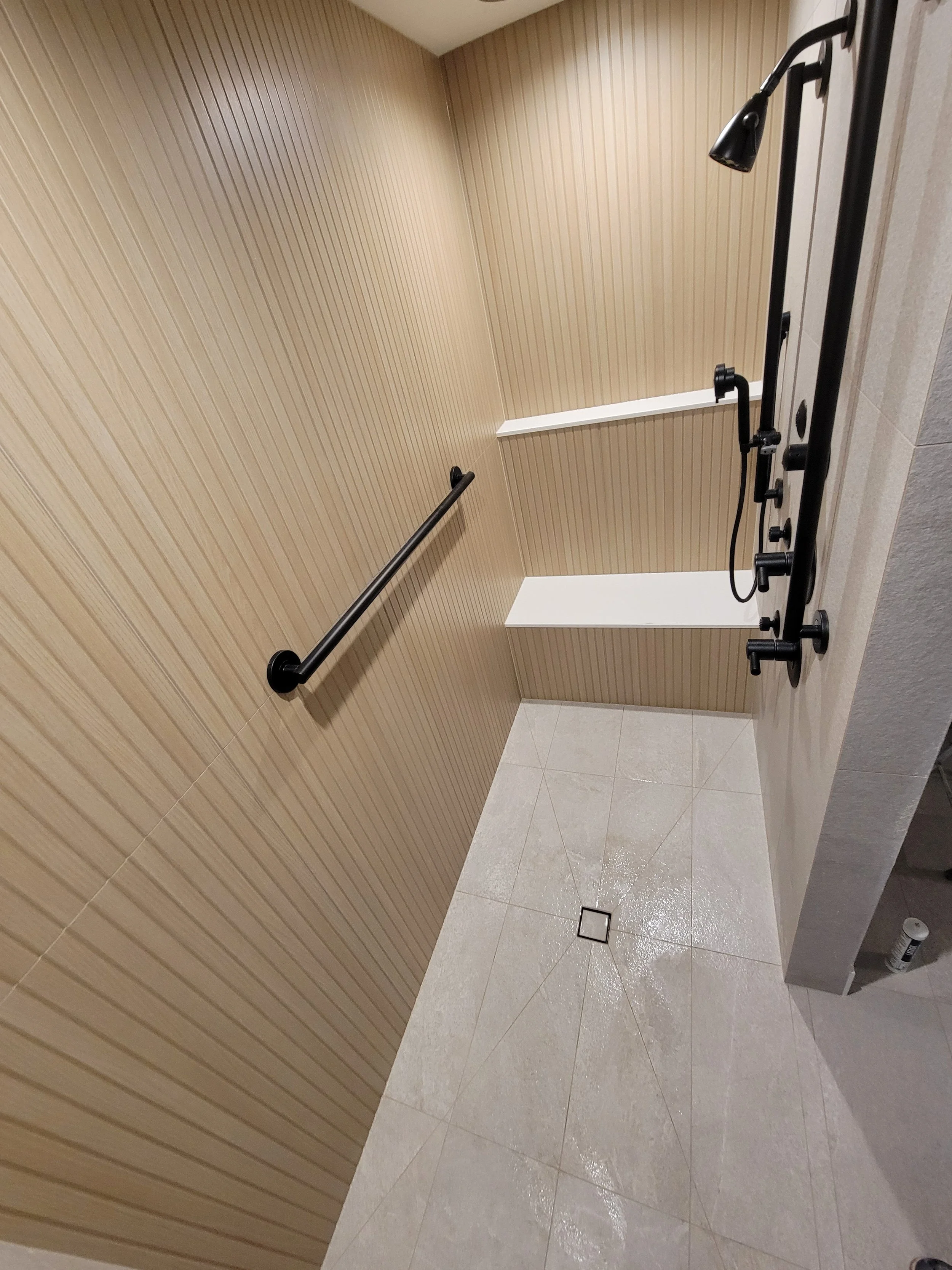 Empty shower area with beige wood-paneled walls, a black grab bar, a black shower fixture with a handheld showerhead, and gray tiled flooring with a small drain.