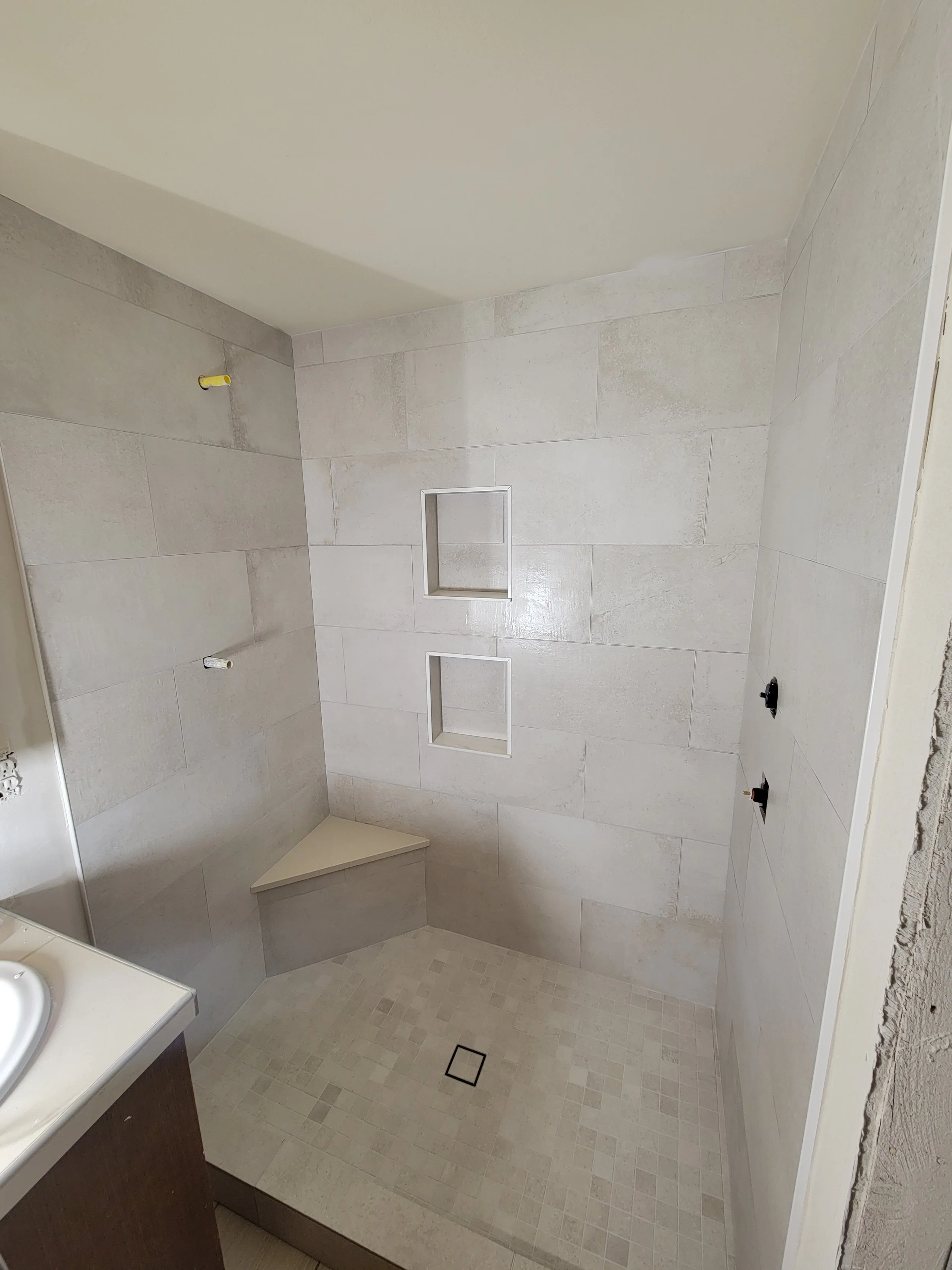 A shower area under construction with two built-in wall niches, a corner bench, and wall fixtures, with the floor covered in small square beige tiles.
