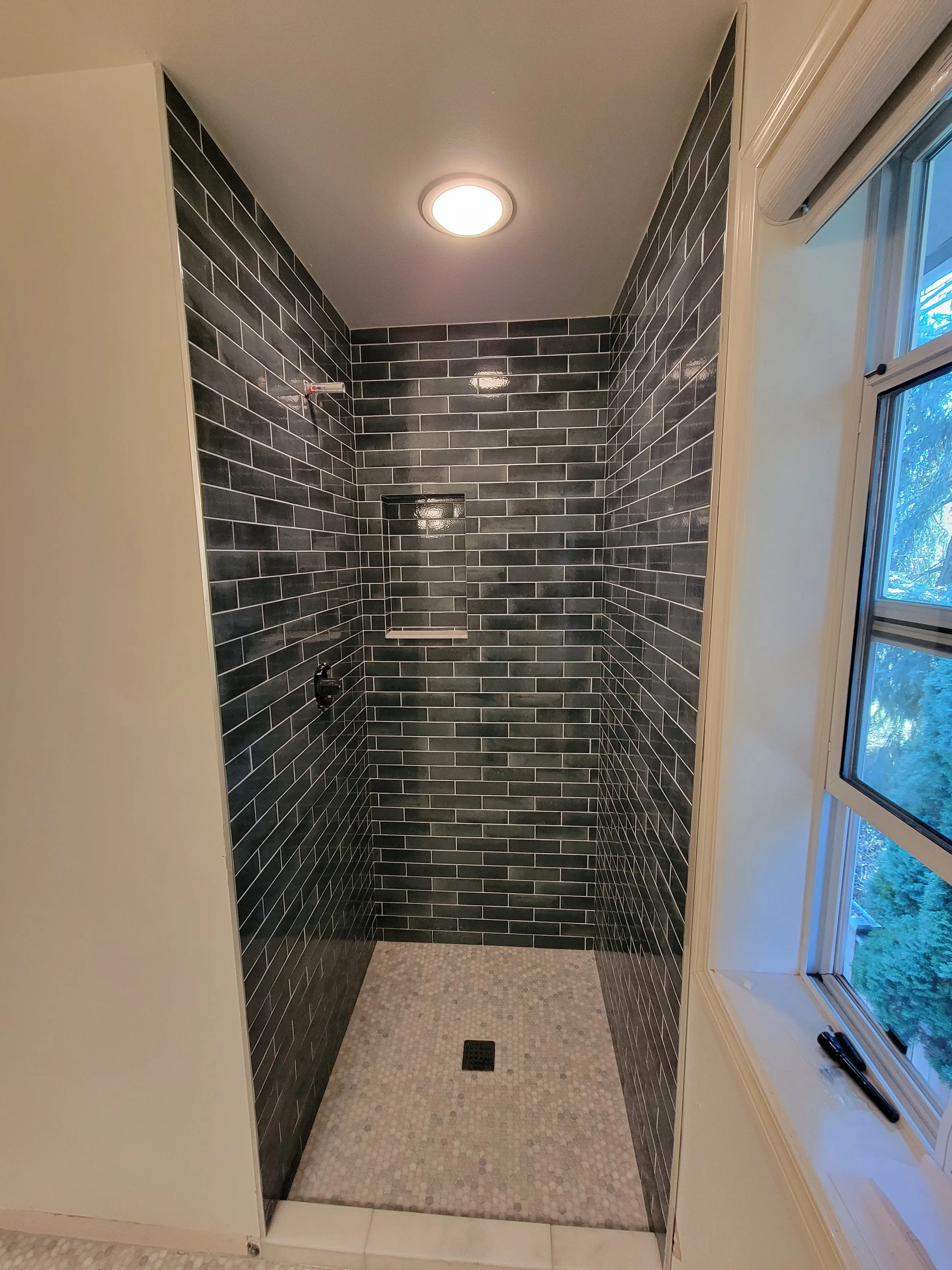 A private shower stall with black subway tile walls, a small niche, and a floor tiled with small light-colored mosaic tiles, next to a window.
