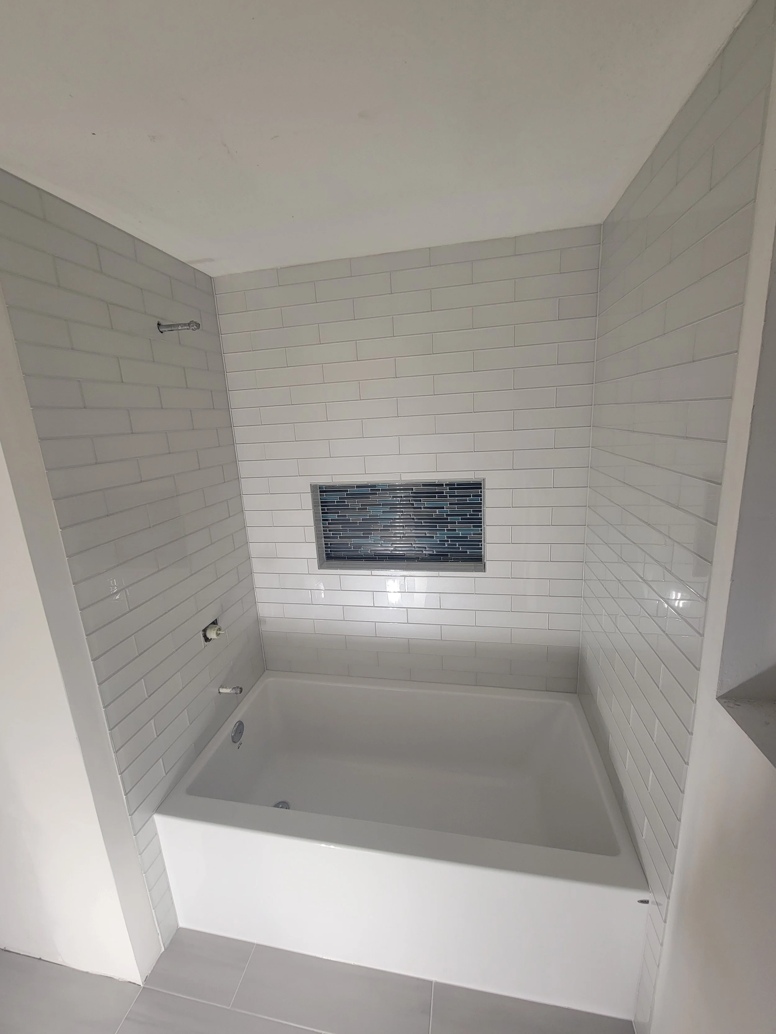 Bathroom bathtub with white subway tile walls, a small window with dark glass tiles, and a gray tiled floor.