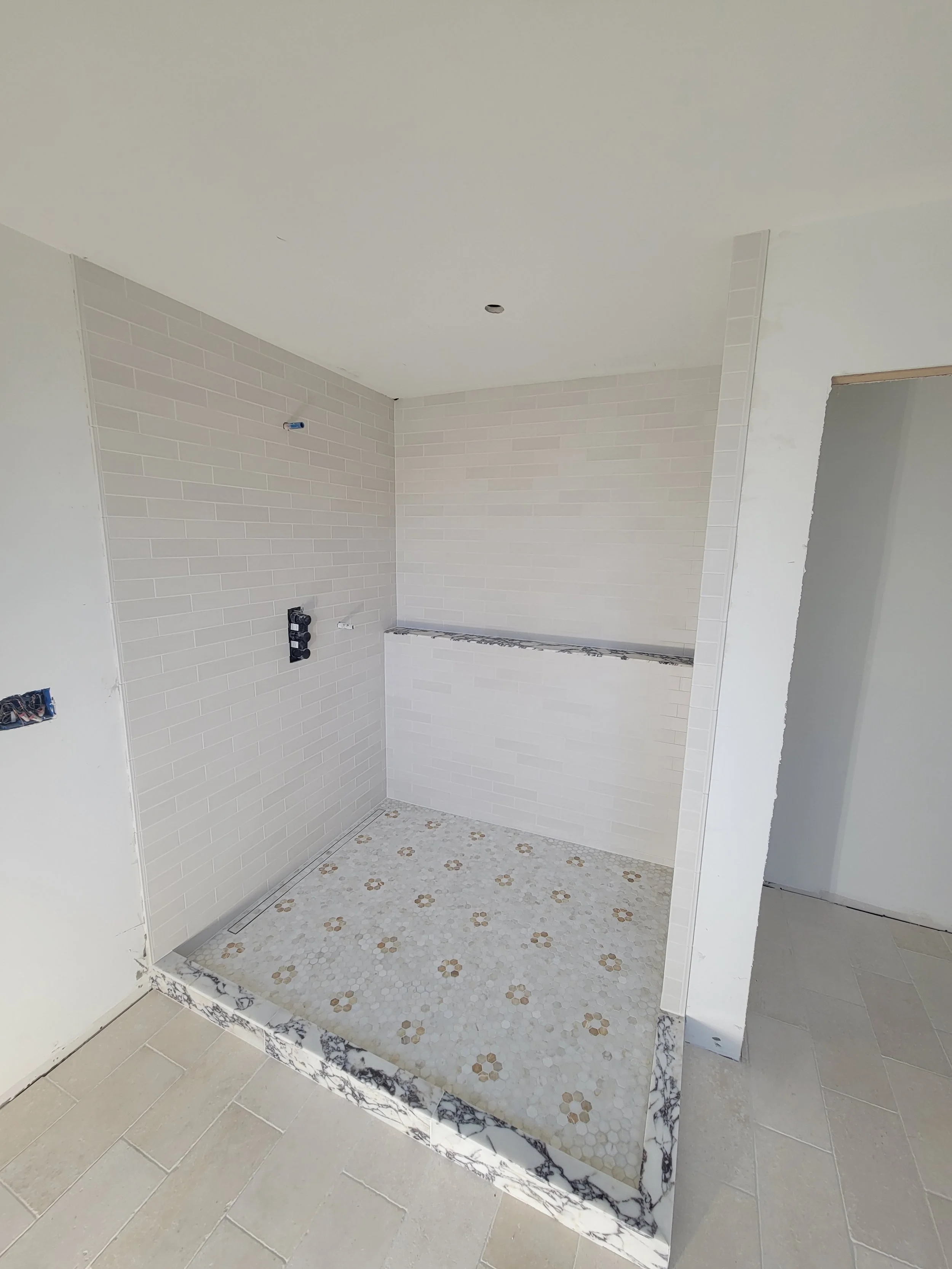 A partially finished shower area with white subway tiles, decorative tile floor with floral pattern, and a marble threshold, in a construction setting.