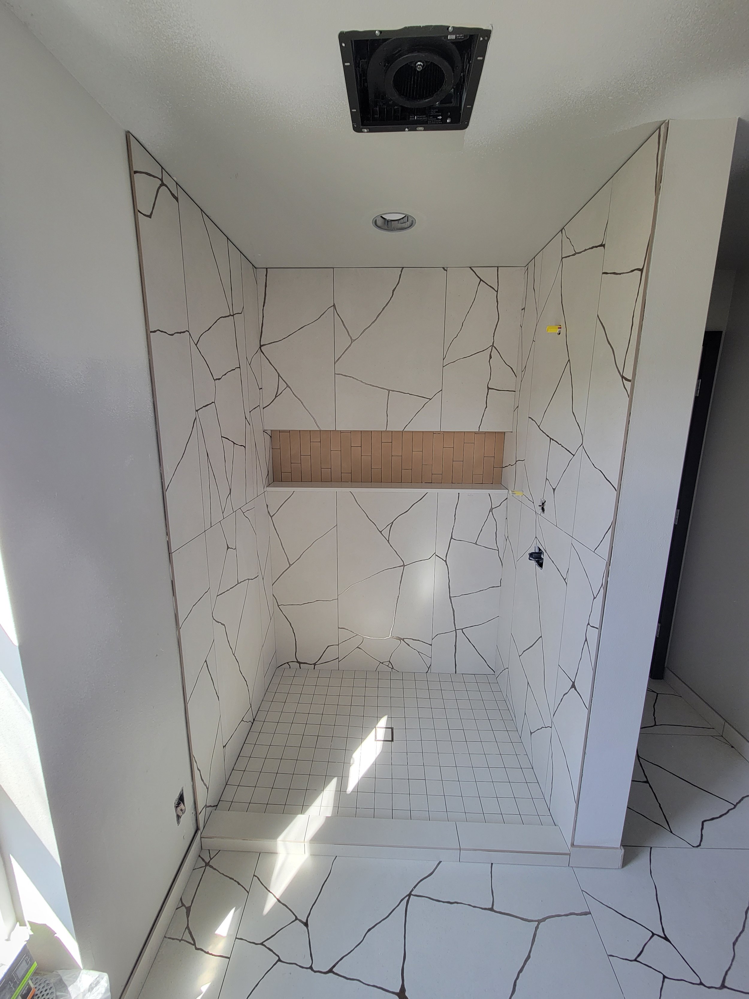 Empty shower in a bathroom with large geometric patterned tiles on the walls and smaller grid tiles on the shower floor, featuring a built-in shelf with brown tiles and an unfinished ceiling with an exhaust fan.