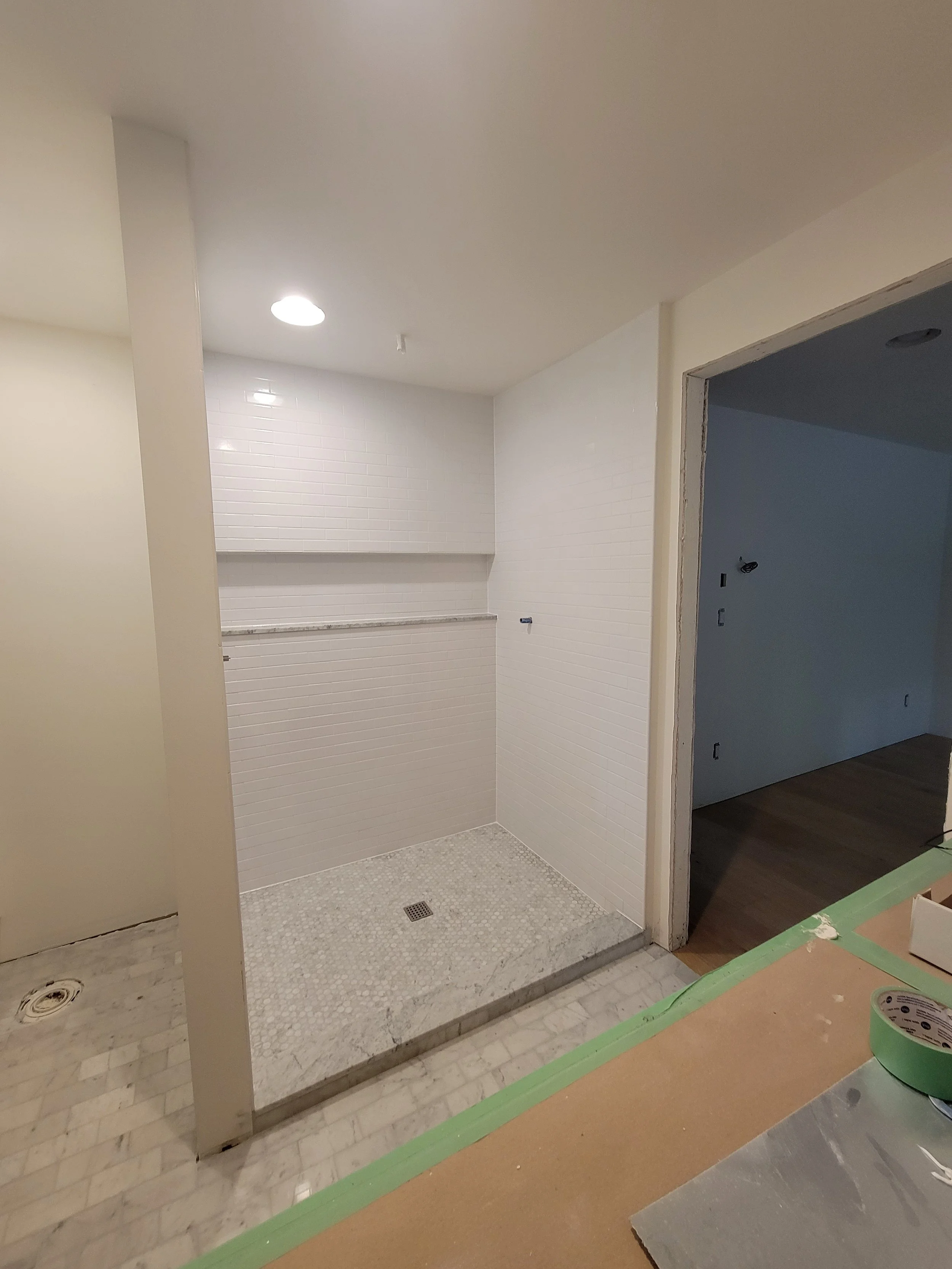A bathroom shower area under construction with white tiles, a small built-in shelf, and a drain on the floor, adjacent to a room with wooden flooring.