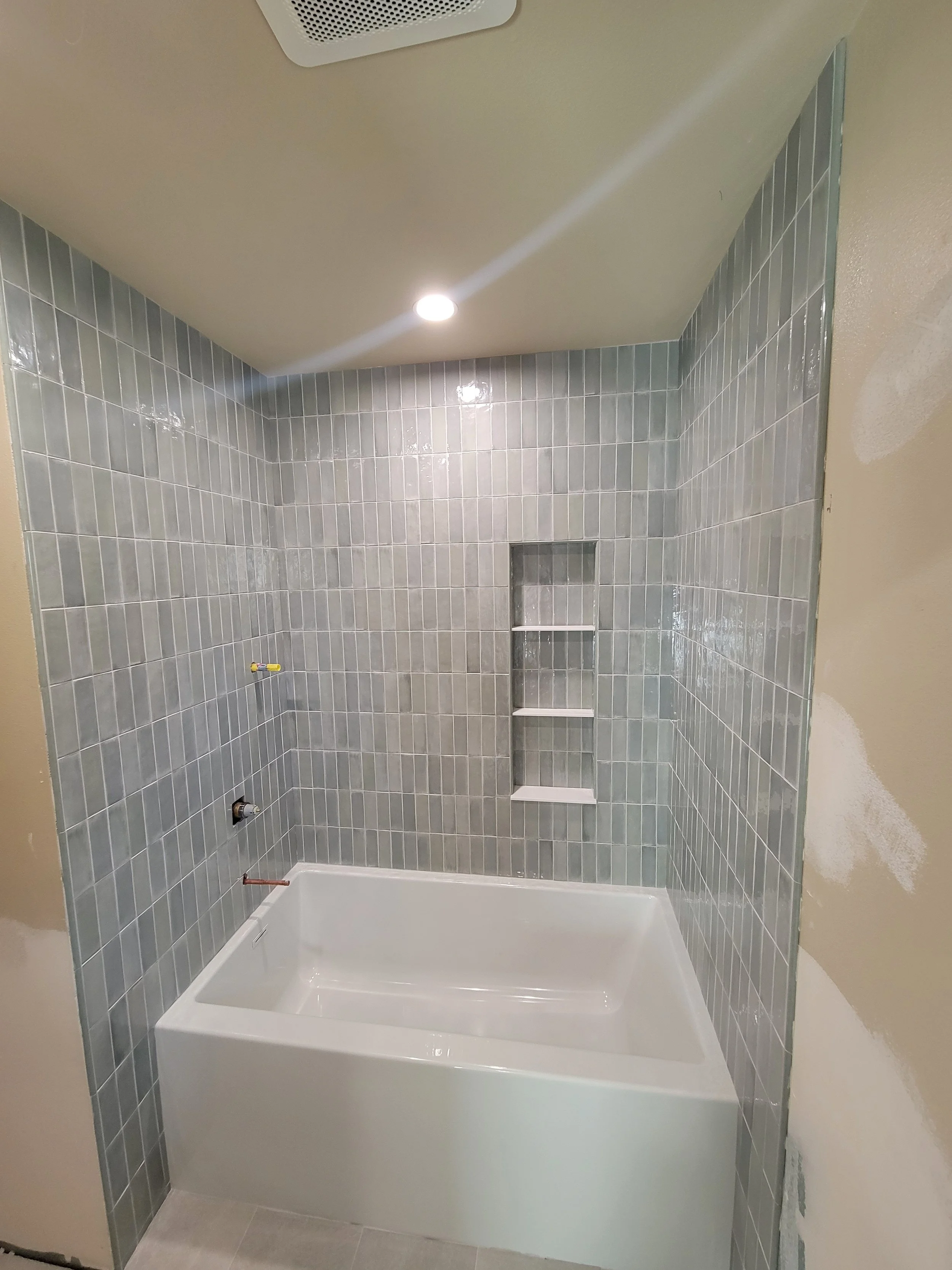 Bathroom with gray tiled walls, white bathtub, built-in shelves, and wiring for fixtures, under construction or renovation.