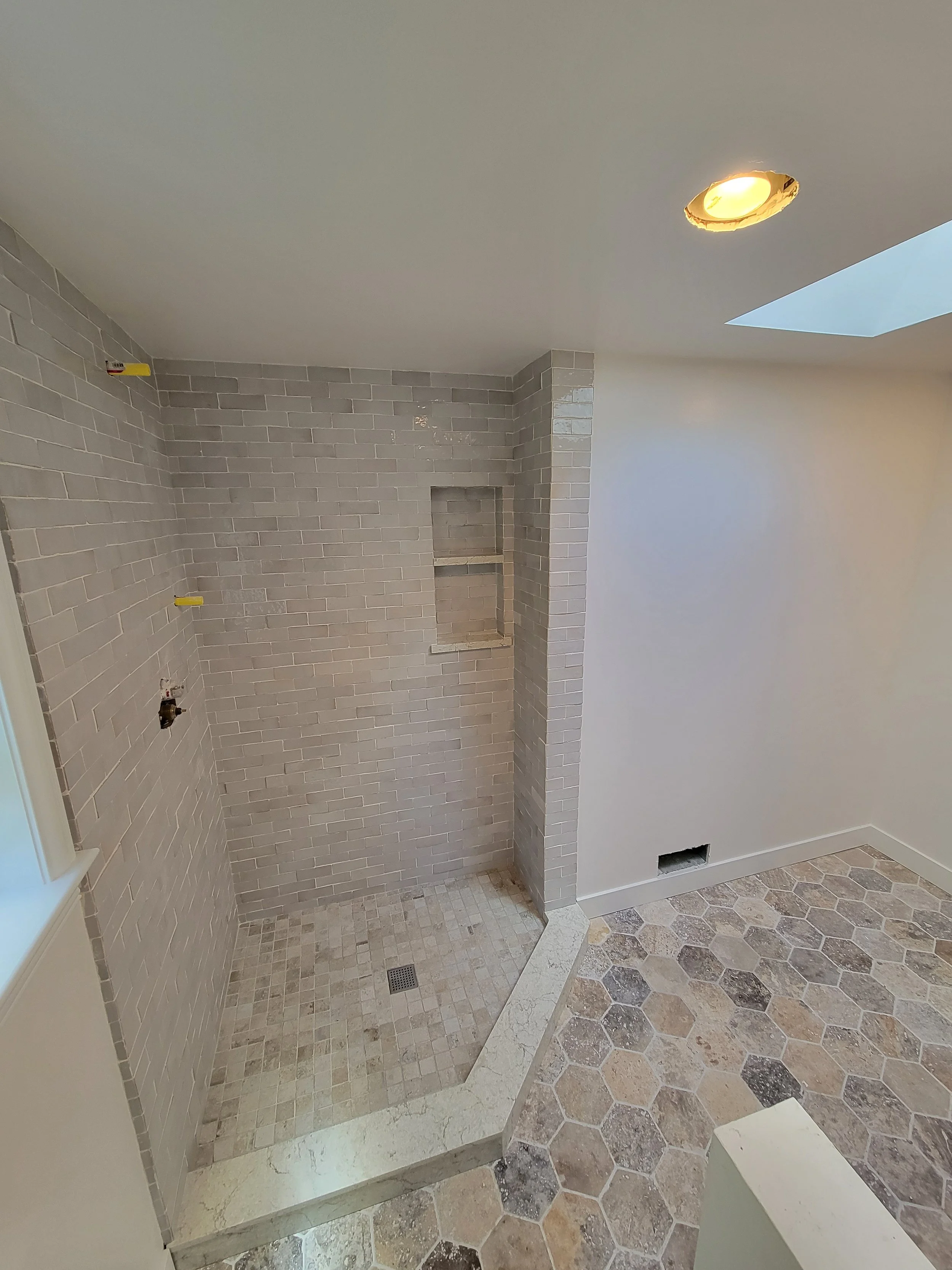 An unfinished bathroom shower with light gray brick tiles, a built-in shelf, and a small tiled floor area. The room has white walls, a hexagonal floor pattern, a ceiling with a round recessed light, and a skylight.