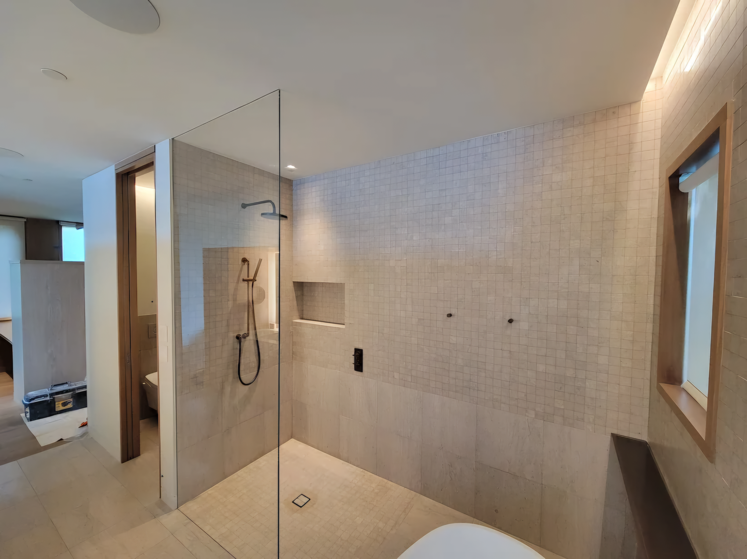 Empty modern walk-in shower with glass partition, beige tiled walls, built-in shelf, and ceiling rain shower.