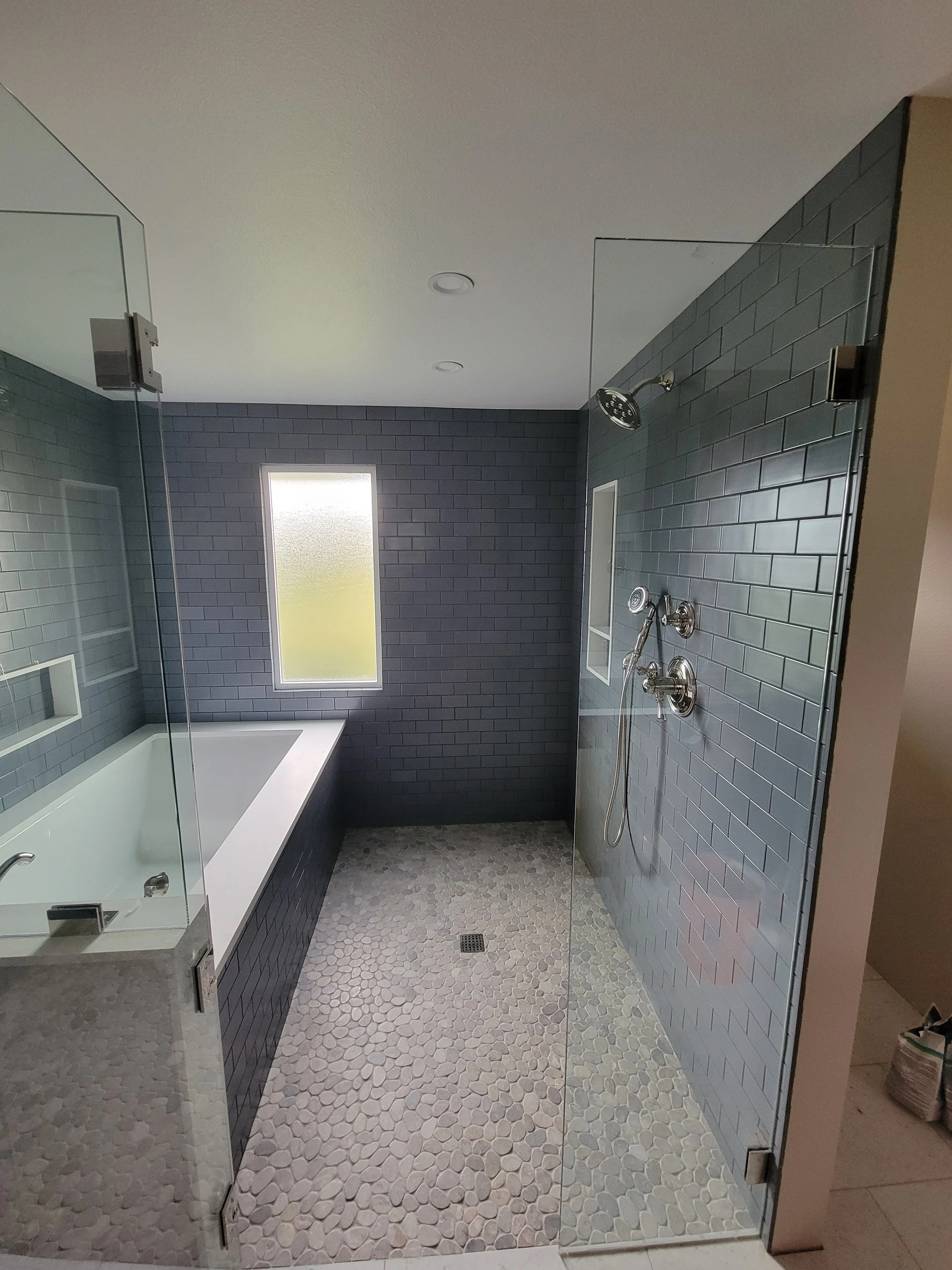 Modern bathroom with pebble stone shower floor, a glass partition, dark gray subway tile walls, a bathtub, and a small frosted window.
