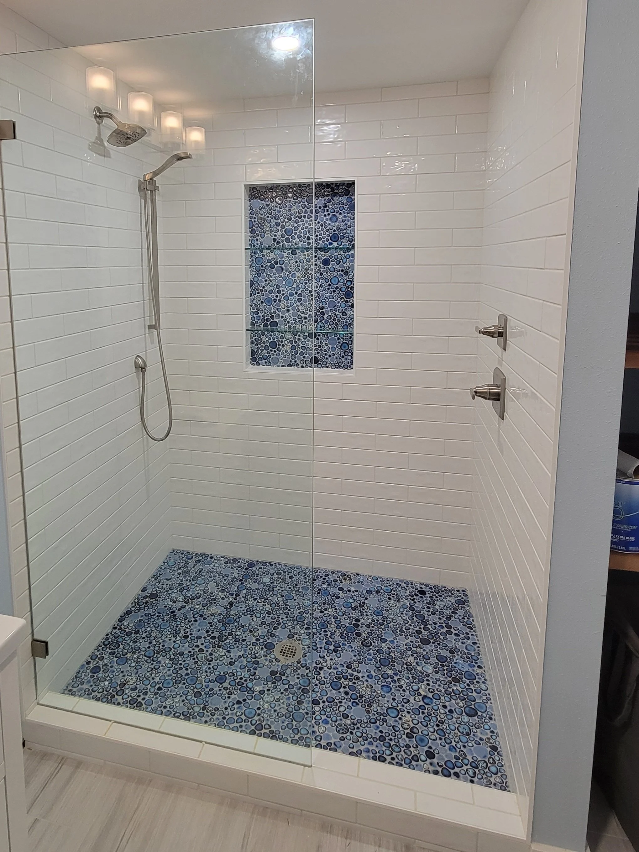 Modern walk-in shower with white subway tile walls, a small window with blue mosaic tiles, a pebble-textured blue tile floor, and brushed nickel fixtures.