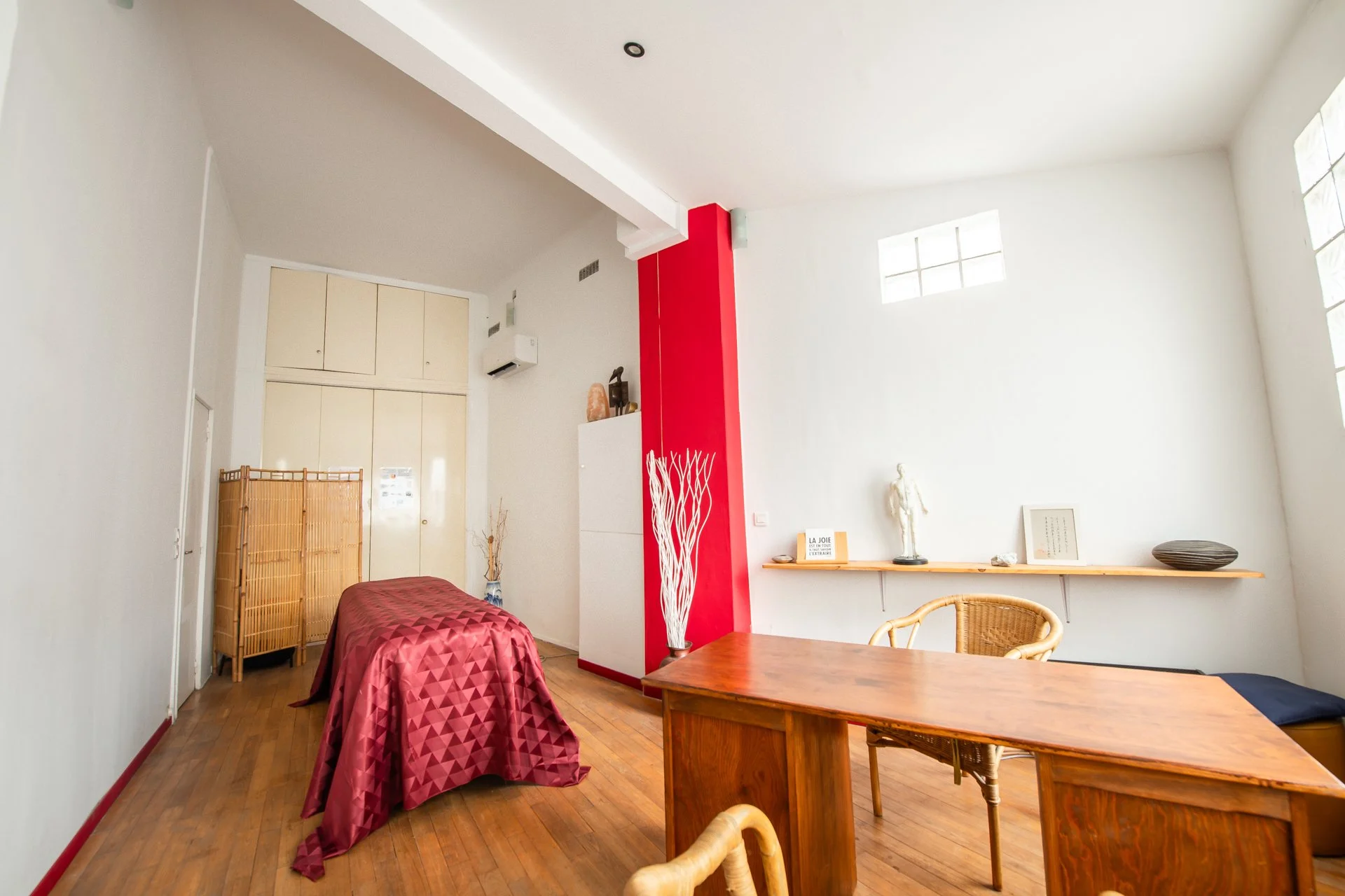 Intérieur lumineux avec un bureau en bois, une chaise en osier, une étagère avec des décorations, et un lit avec une couverture rouge. Mur blanc avec une fenêtre en parpaings de verre.