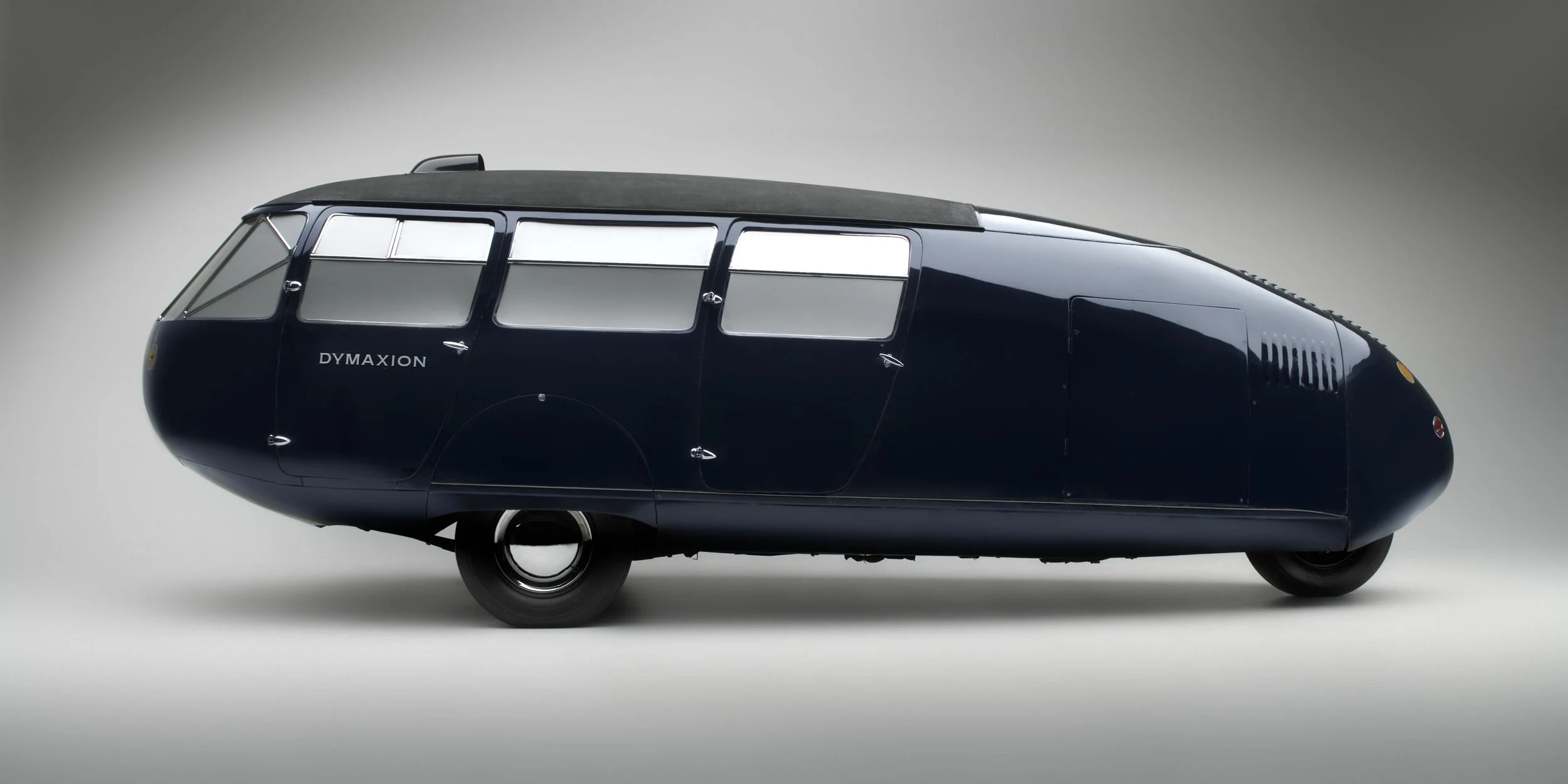 A sleek black vintage Dymaxion car designed by Buckminster Fuller, a curved, streamlined design and multiple windows, set against a plain light gray background.