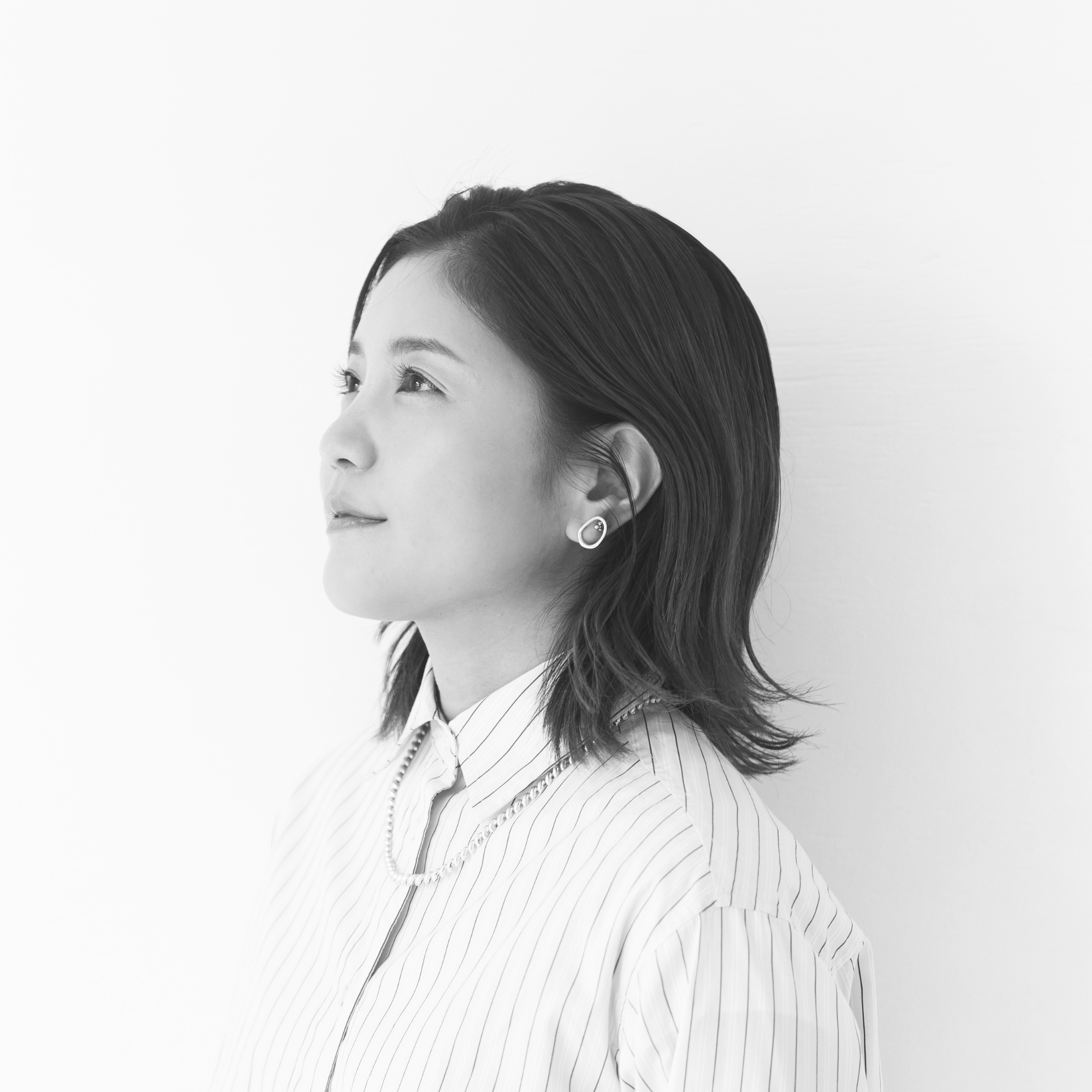 Black and white photo of a woman with shoulder-length straight hair, wearing a striped shirt, earrings, and a necklace, looking to the left with a slight smile.