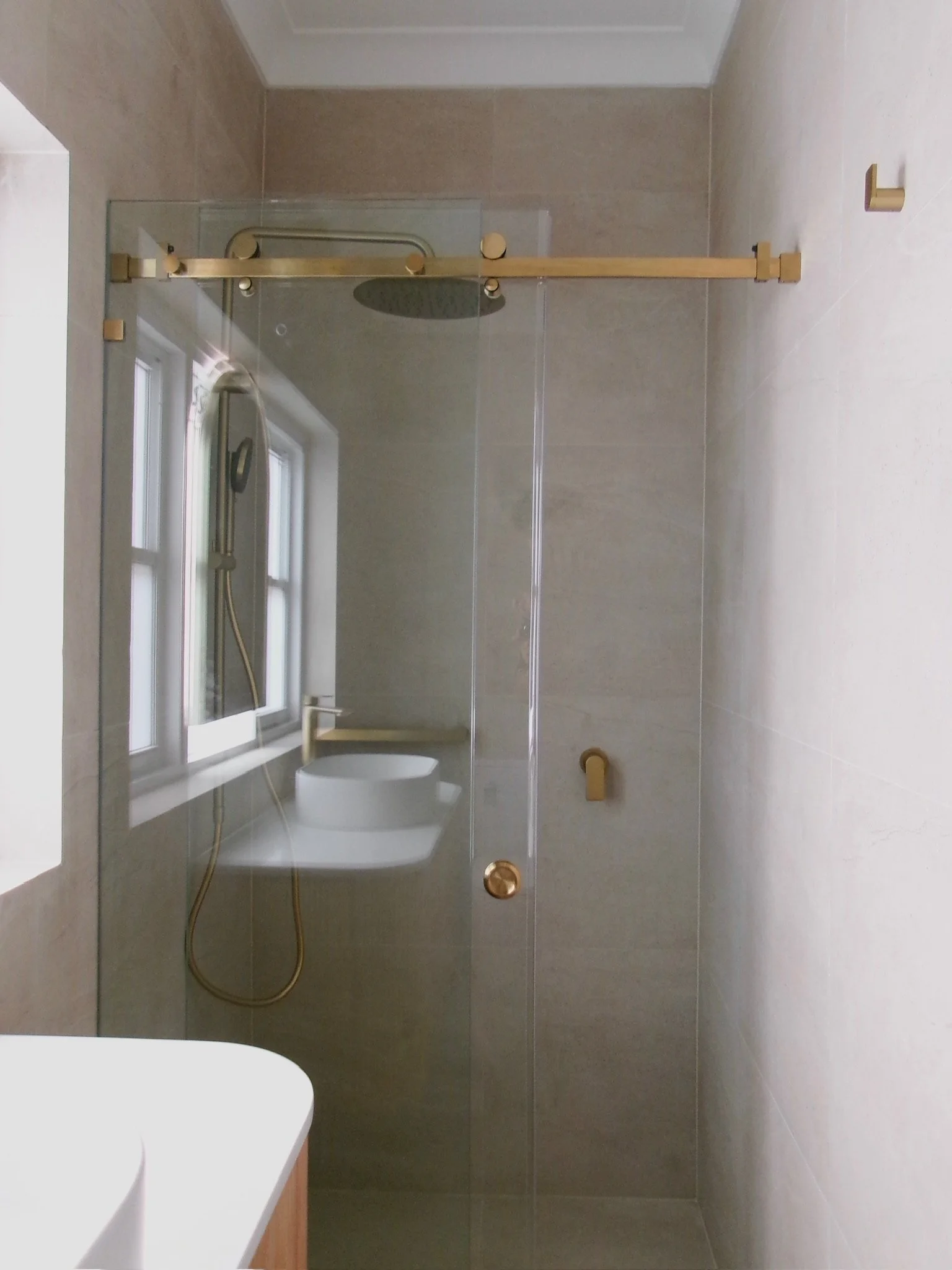 Modern bathroom with shower area enclosed by a glass door, a large rain showerhead, a hand-held shower, a small window, and minimalist fixtures in gold finish.