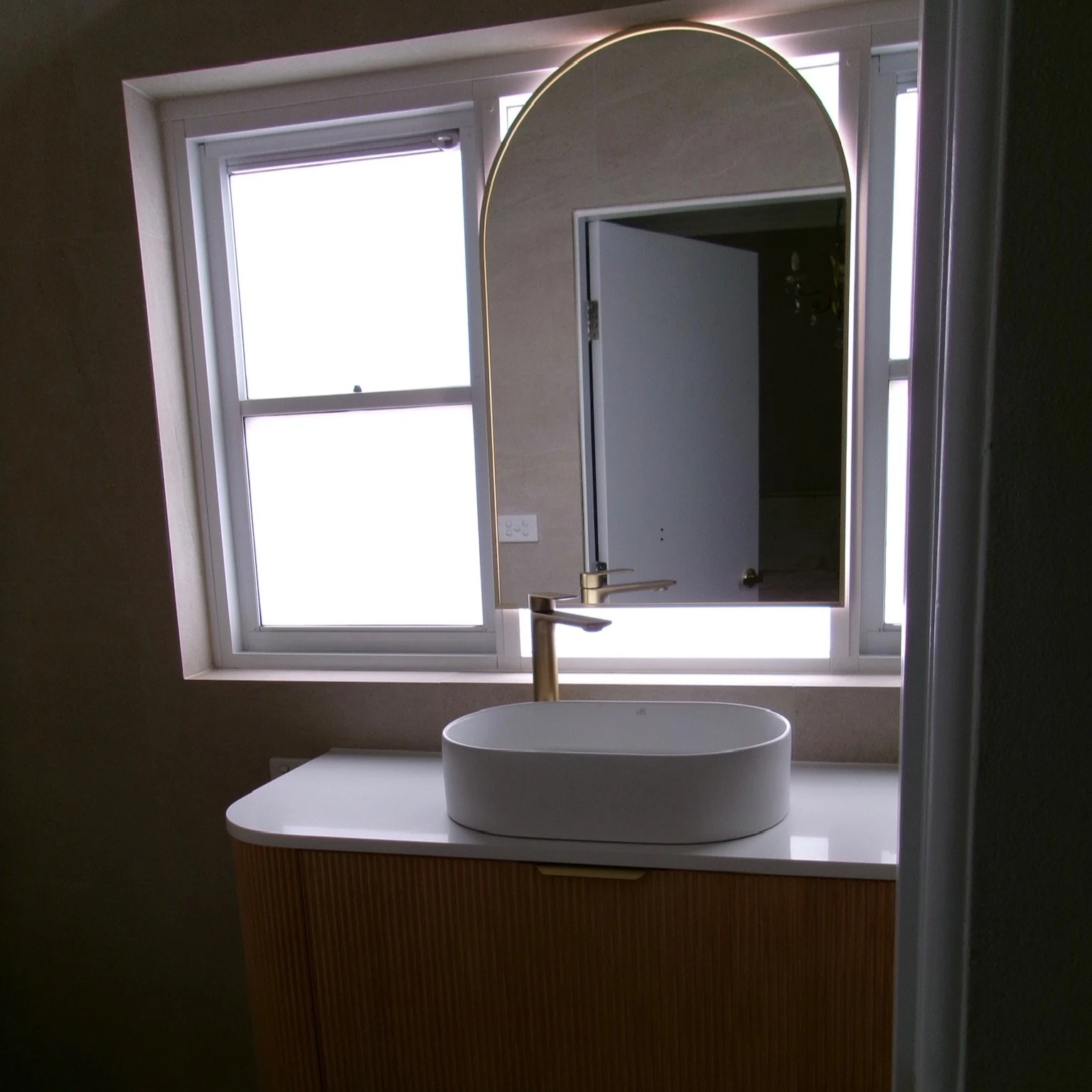 Modern bathroom vanity with a white oval vessel sink and a gold faucet, positioned beneath a mirror with an arched top and a window in the background.