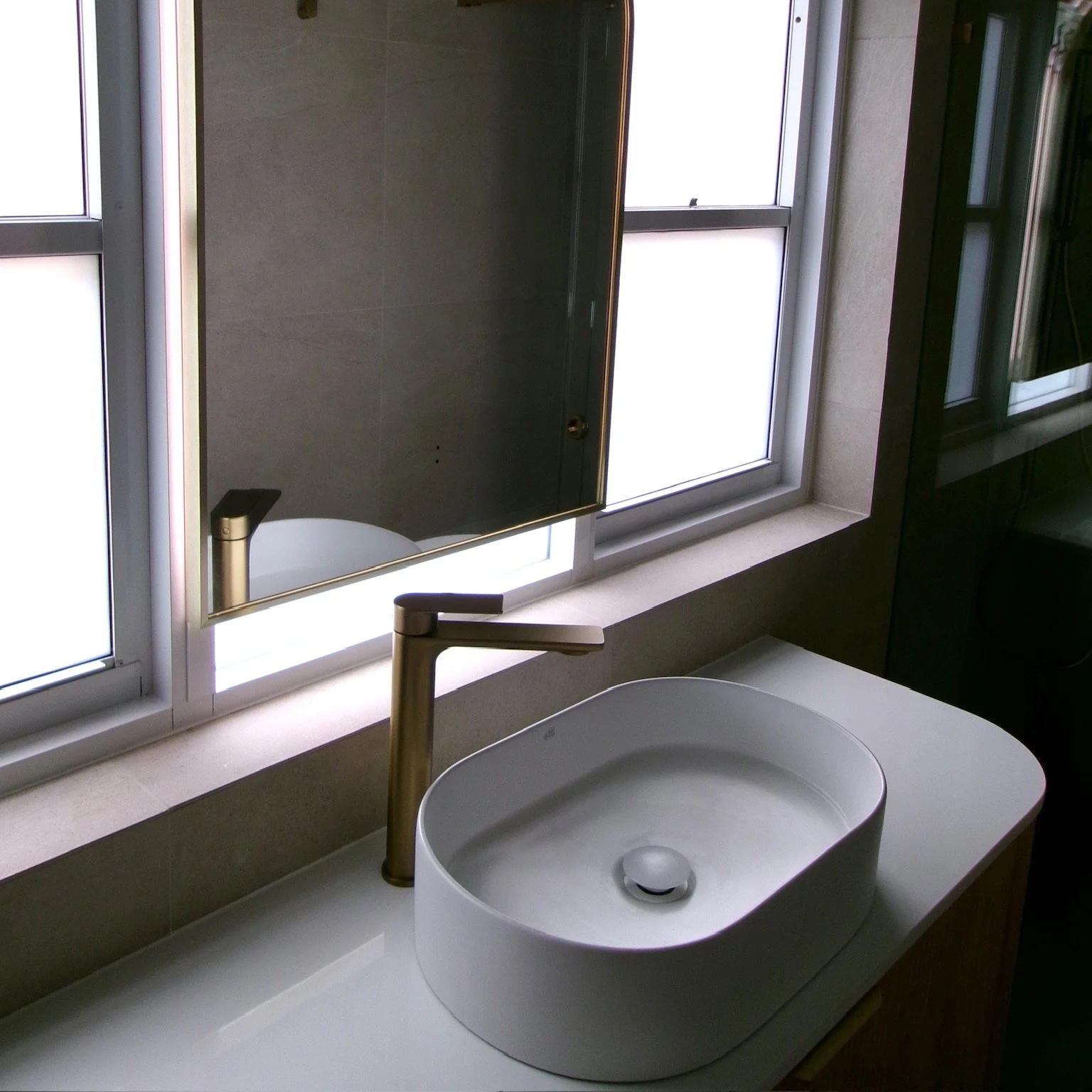 Bathroom countertop with a white vessel sink and a gold faucet, above a large window with a mirror reflecting the sink.
