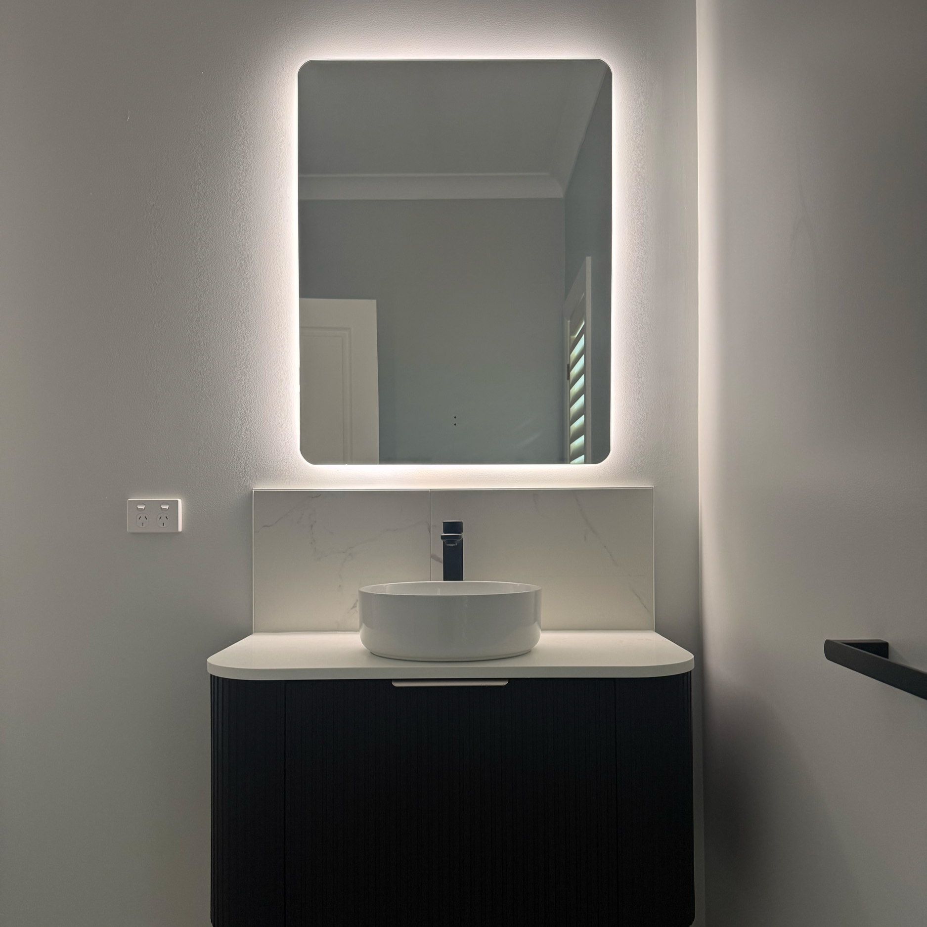 Modern bathroom vanity with a round vessel sink, black faucet, and a backlit square mirror on a white wall.