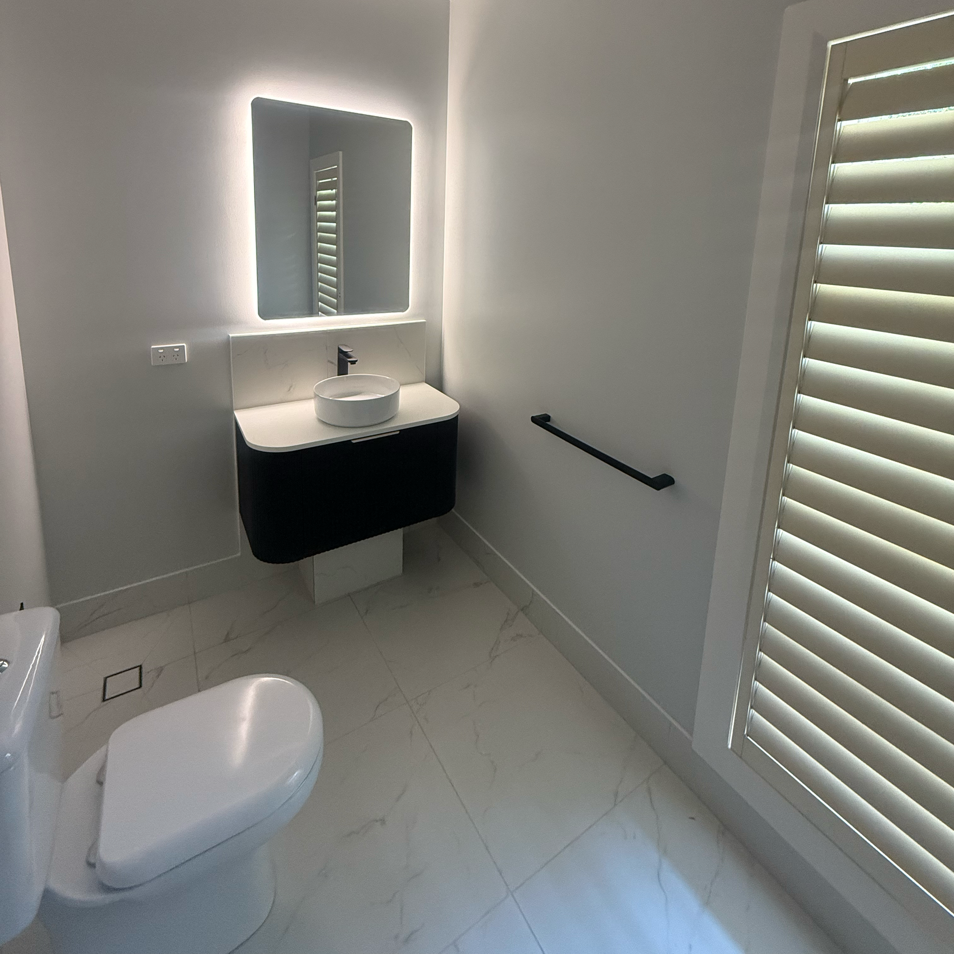 A modern bathroom with white walls, marble flooring, a black floating vanity with a round white vessel sink, a large illuminated mirror, a window with white shutters, a black handrail on the wall, and a nearby toilet.