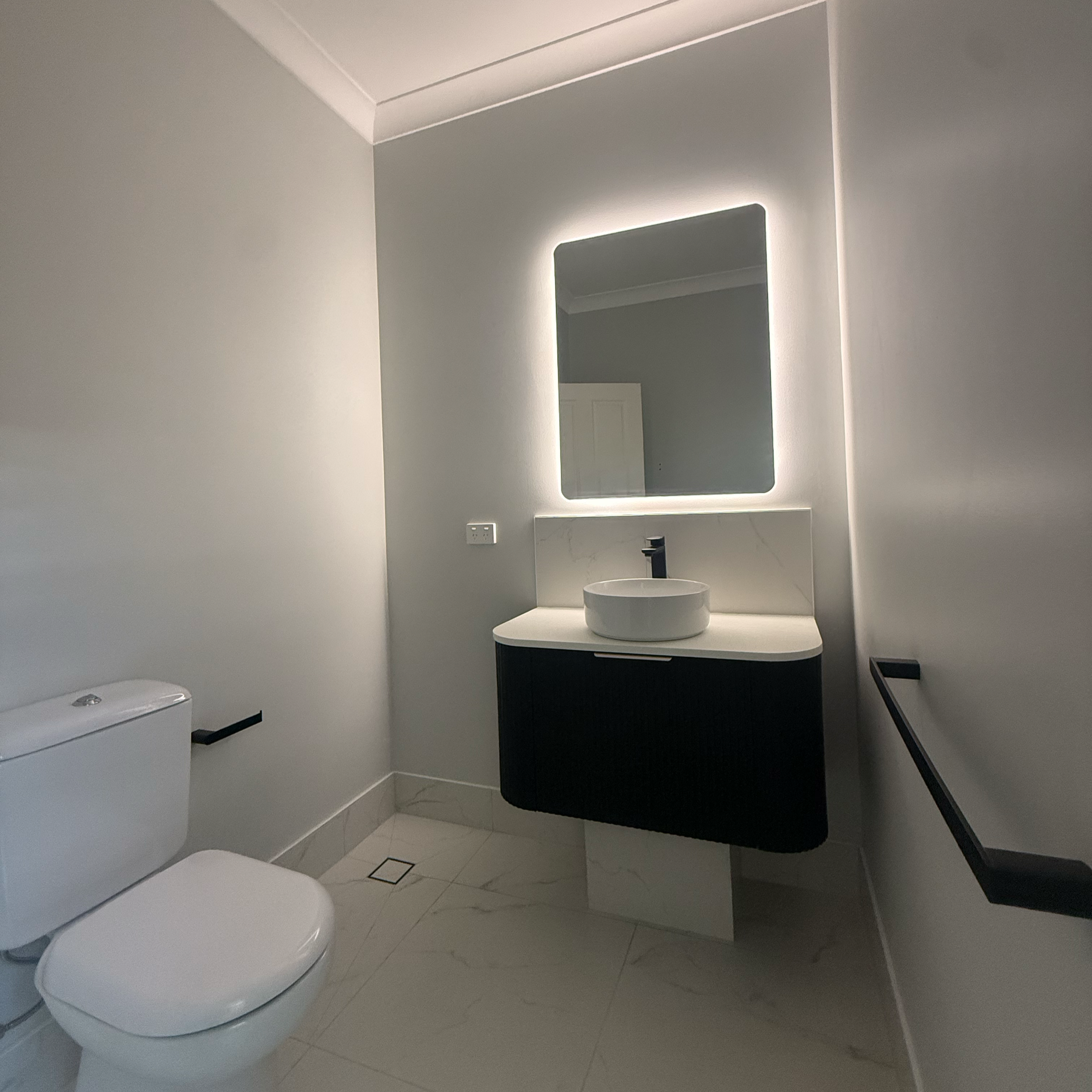 Modern bathroom with white toilet, black and white vanity with round basin sink, illuminated rectangular mirror, white marble floor, and gray walls.