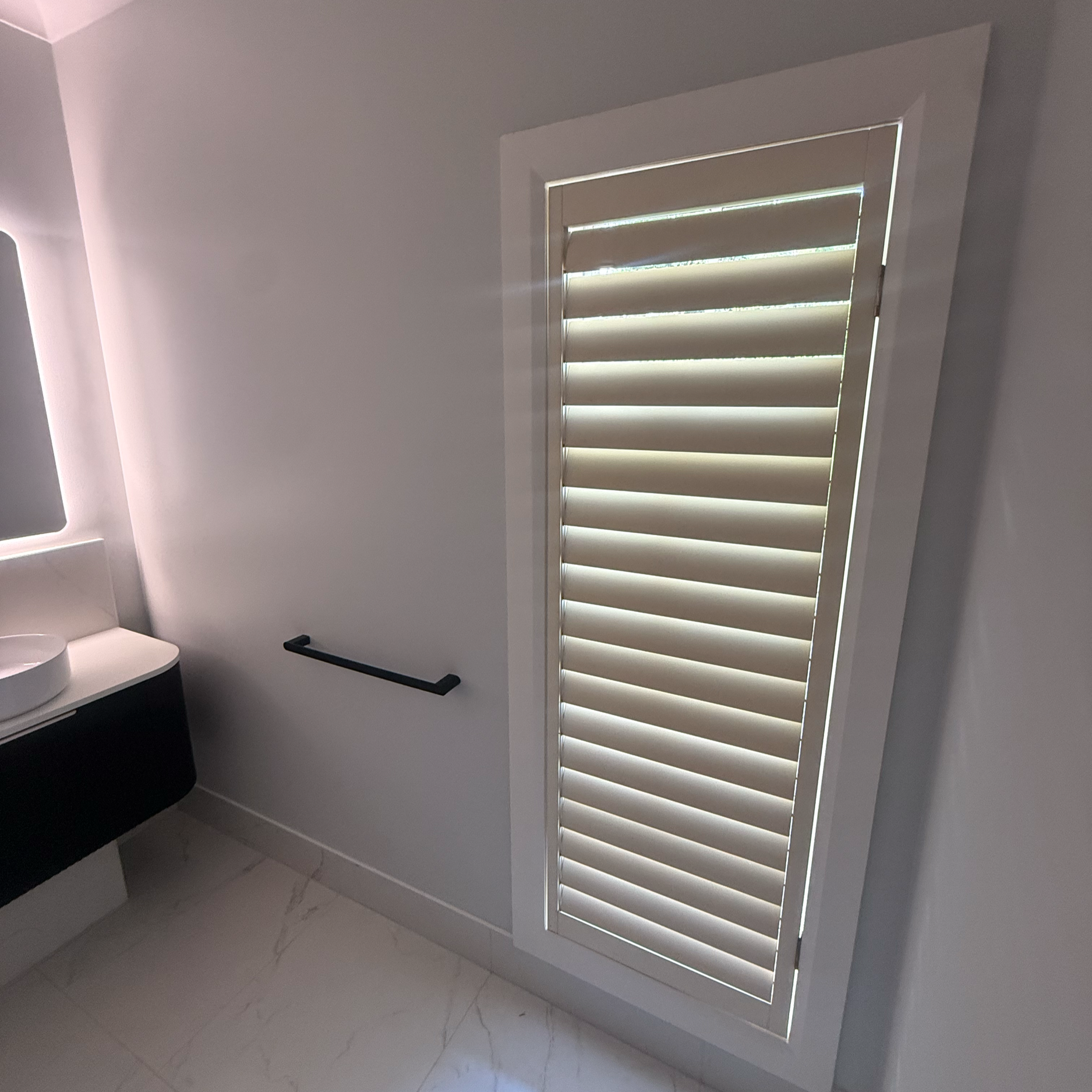 A bathroom with a white wall, a window with white plantation shutters, a black towel rail, and a white sink with a black cabinet.