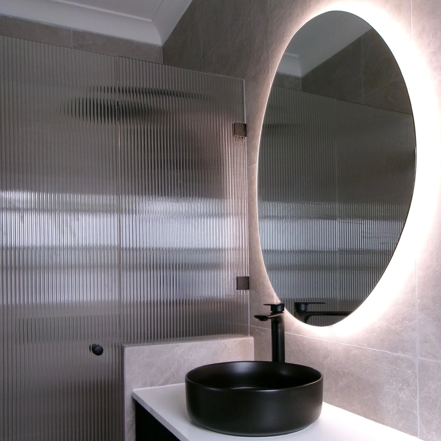 Modern bathroom with a black vessel sink, a large backlit mirror, and textured frosted glass partition.