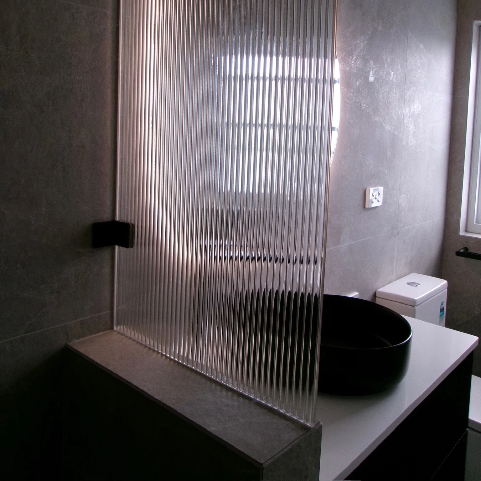 Bathroom with a textured glass partition, a round black vessel sink on a white countertop, a white toilet, and a window.
