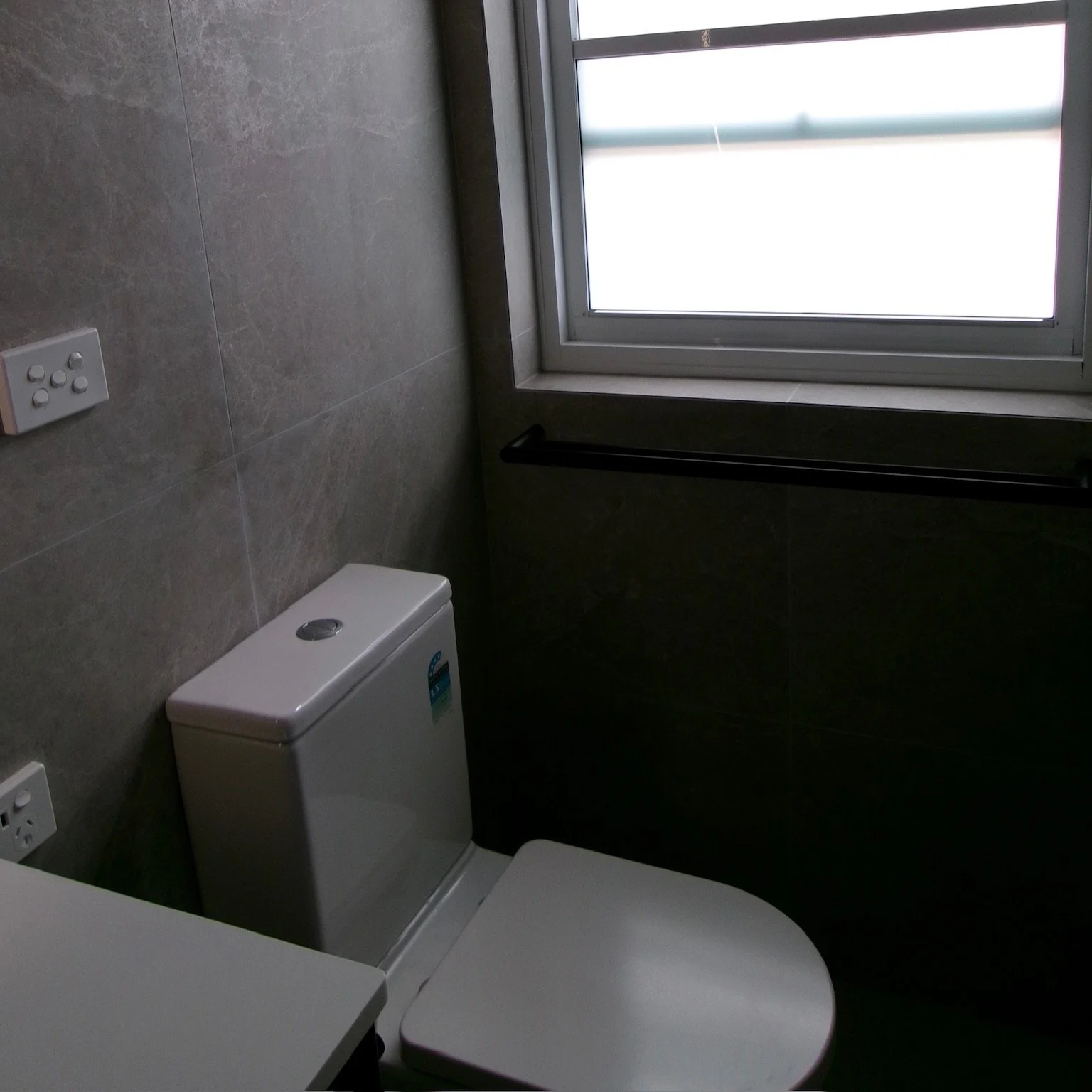A small bathroom with tiled walls, a white toilet, a window allowing natural light, and an electrical outlet on the wall.