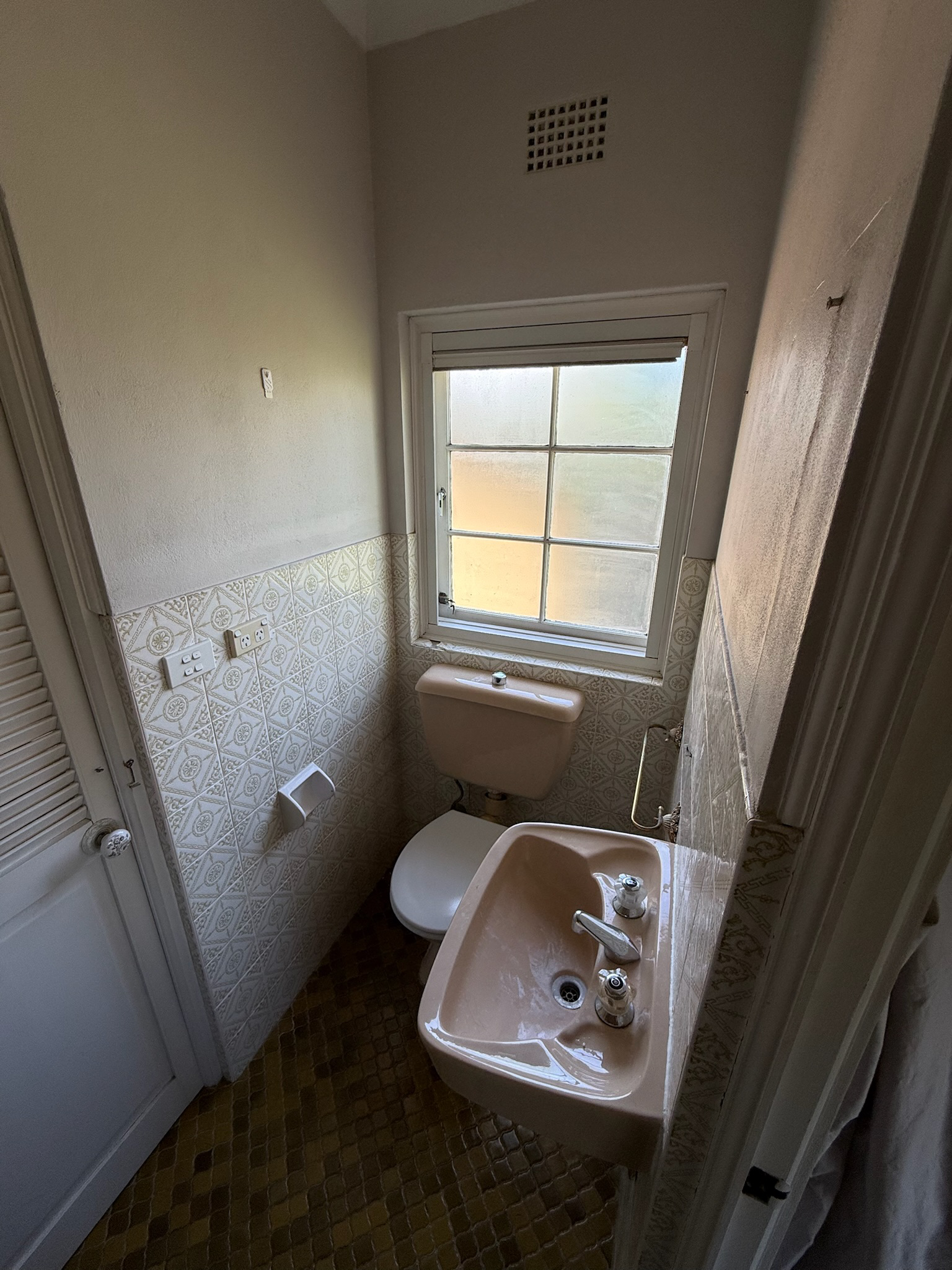 Small vintage bathroom with beige sink, toilet, patterned wall tiles, frosted window, and a ceiling vent.