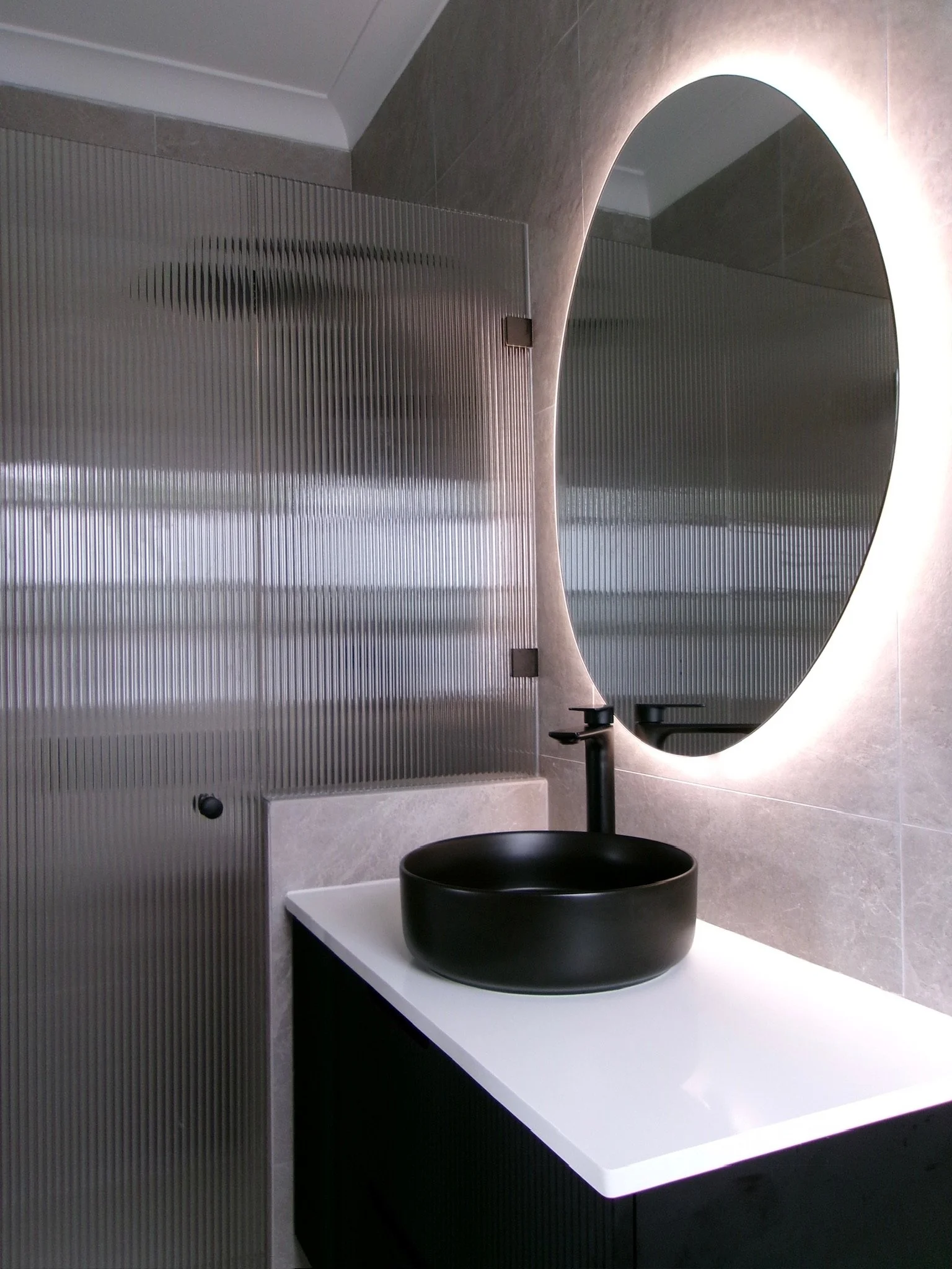 Modern bathroom with a black vessel sink, black faucet, large oval mirror, and textured glass partition.