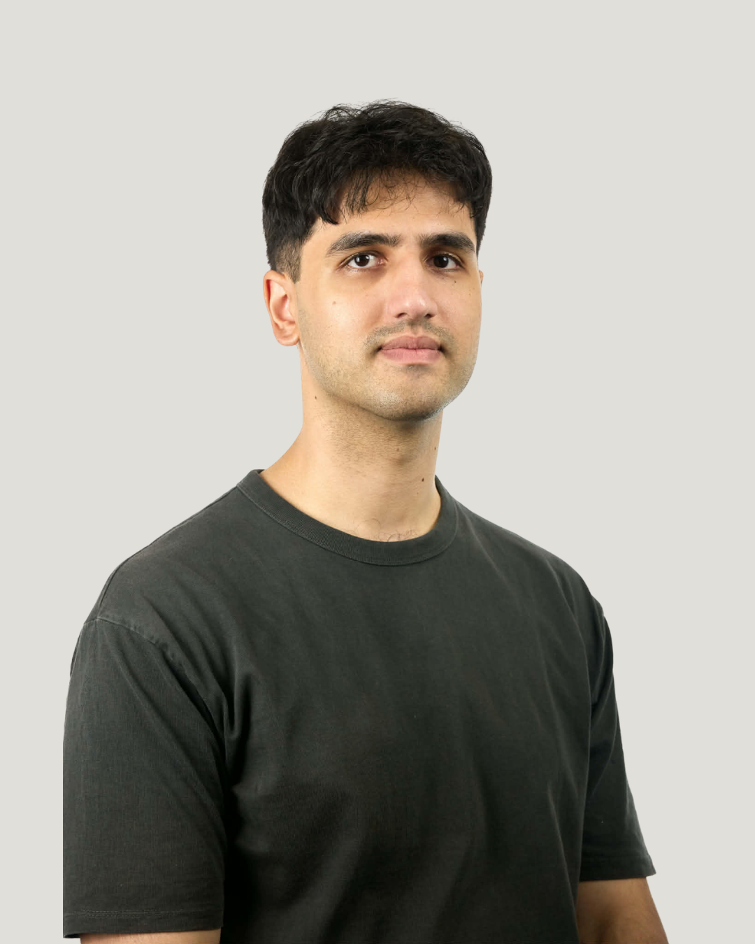 A young man with dark hair, wearing a black t-shirt, looking at the camera against a plain light gray background.