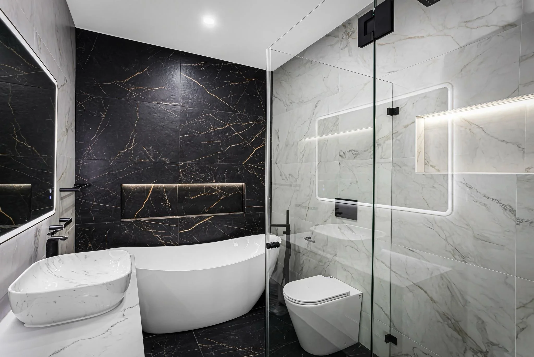 Modern bathroom with black marble accent wall behind a freestanding bathtub, white marble tiles on other walls, a glass shower enclosure with built-in lighting, a wall-mounted toilet, and a marble vessel sink.