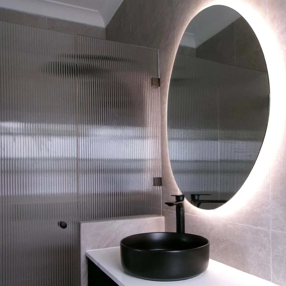 Modern bathroom with a large round mirror, a black vessel sink, and a sleek faucet. The wall features textured, vertical metal panels and smooth beige tiles.