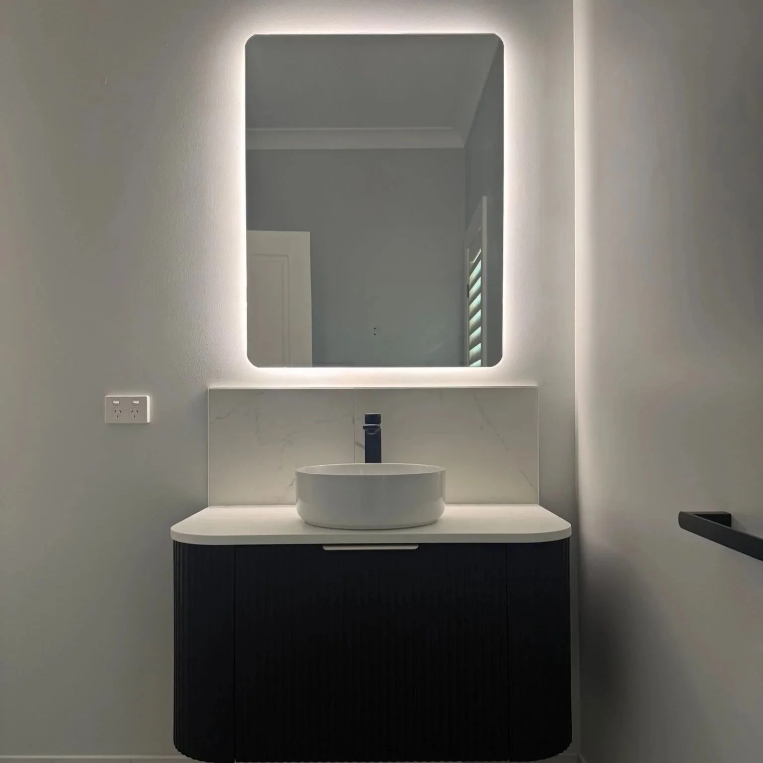 Modern bathroom vanity with a white vessel sink, black faucet, illuminated rectangular mirror, and dark cabinet below, set against light-colored walls.