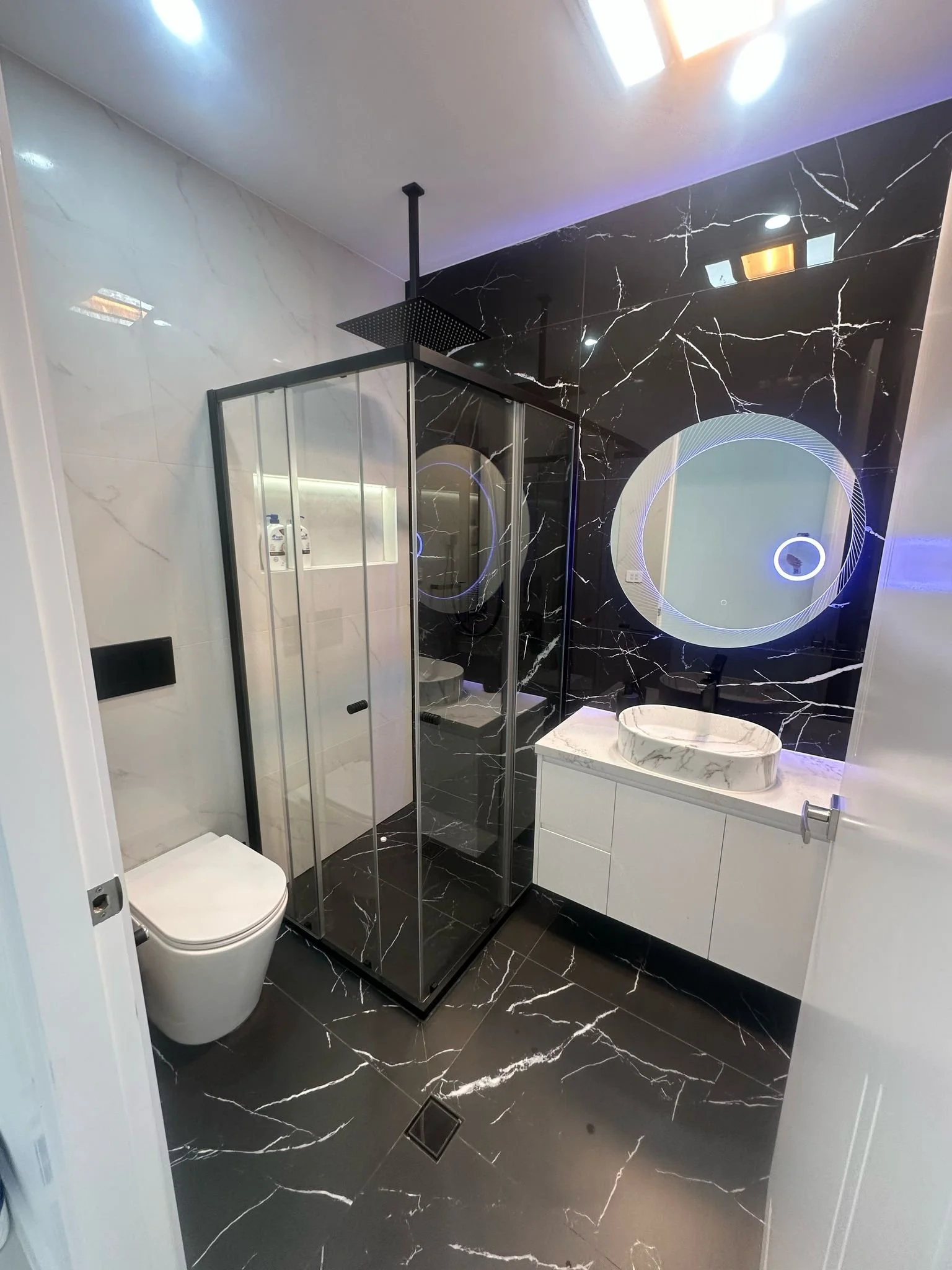 Modern bathroom with black marble wall and floor, glass shower enclosure, white vanity with round marble sink, backlit circular mirror, toilet, and LED ceiling lights.