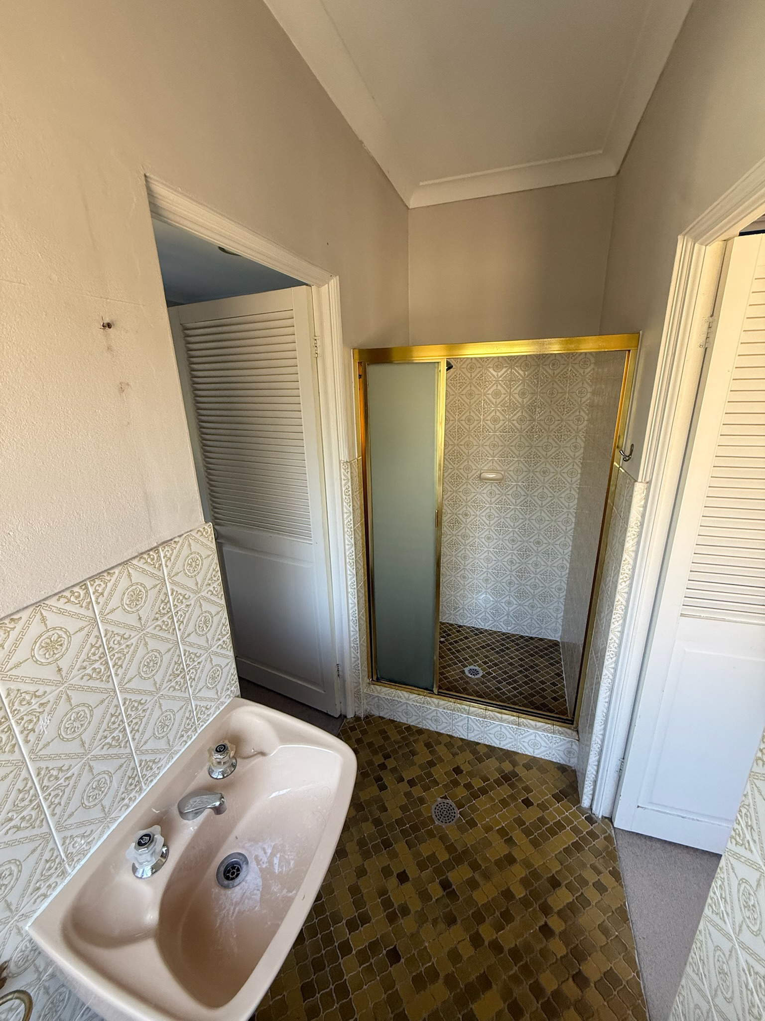 Small bathroom with a beige sink, brown tiled floor, and a shower with gold trim and frosted glass door.