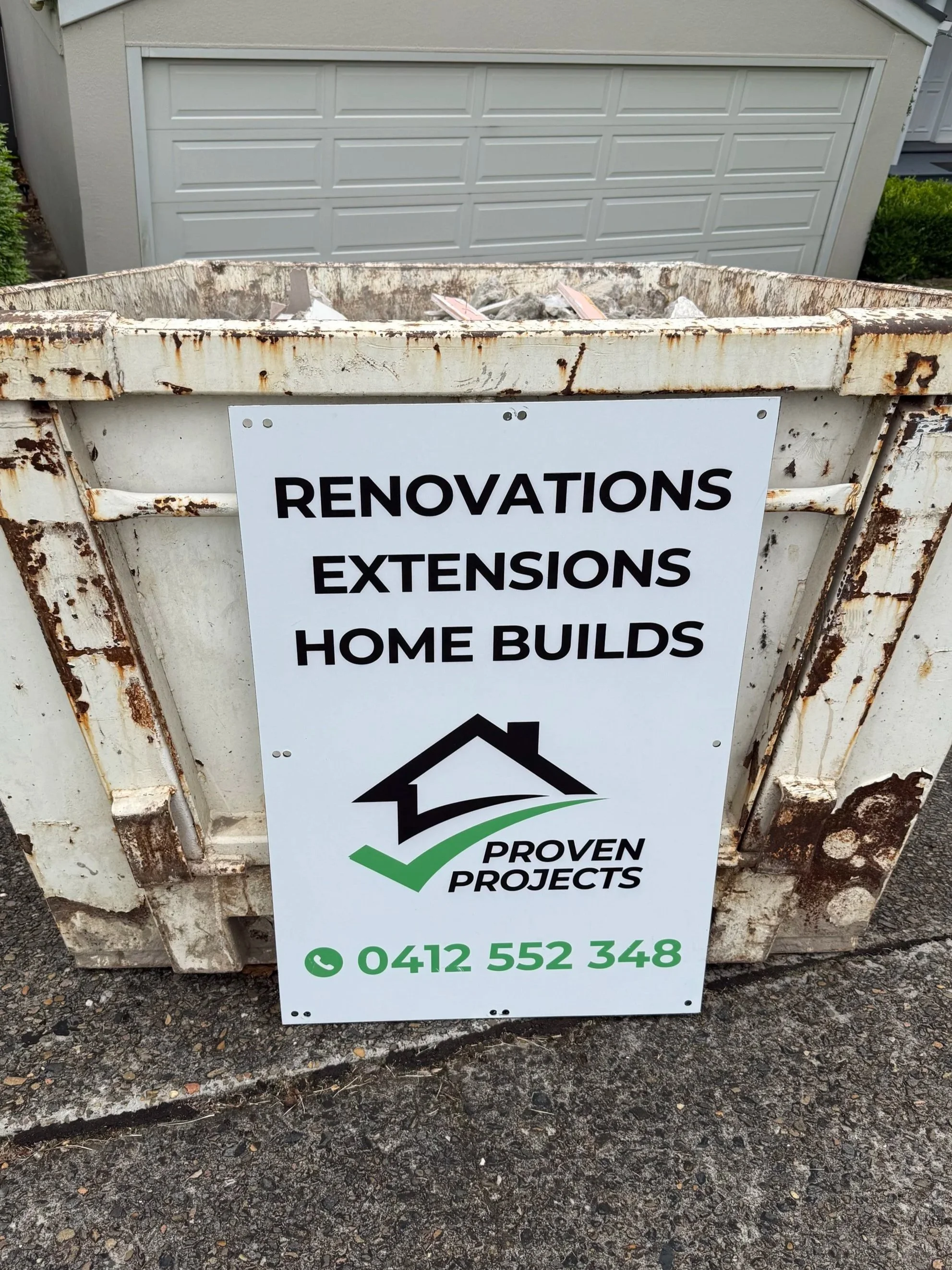 Sign on a rusty construction dumpster advertising renovation, extension, and home build services by Proven Projects, with a house logo and contact number 0412 552 348.
