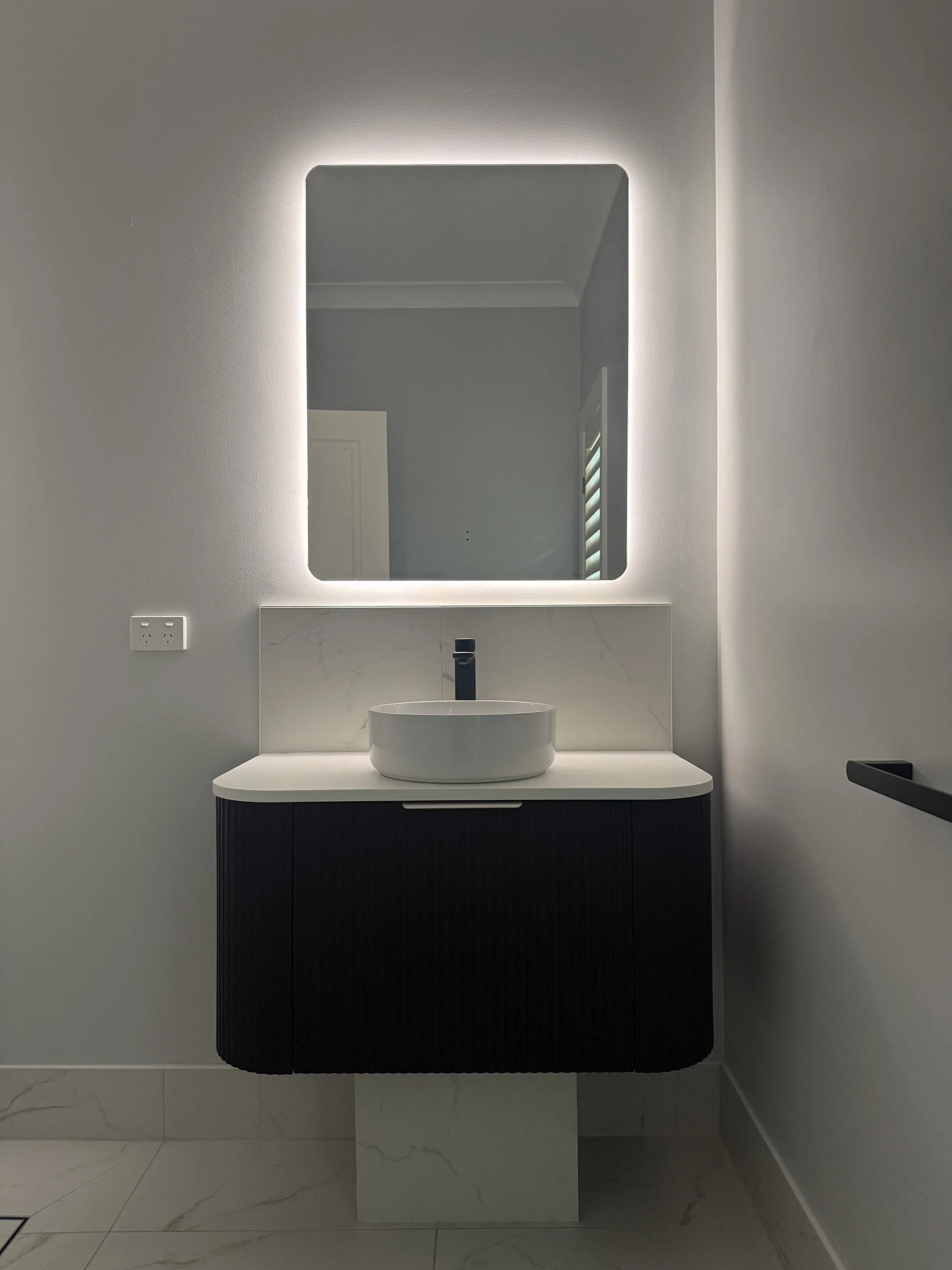 Modern bathroom with a backlit mirror, a white vessel sink, black faucet, and a black vanity with vertical ridges, set against a light gray wall with a white marble backsplash. A window with blinds is reflected in the mirror.