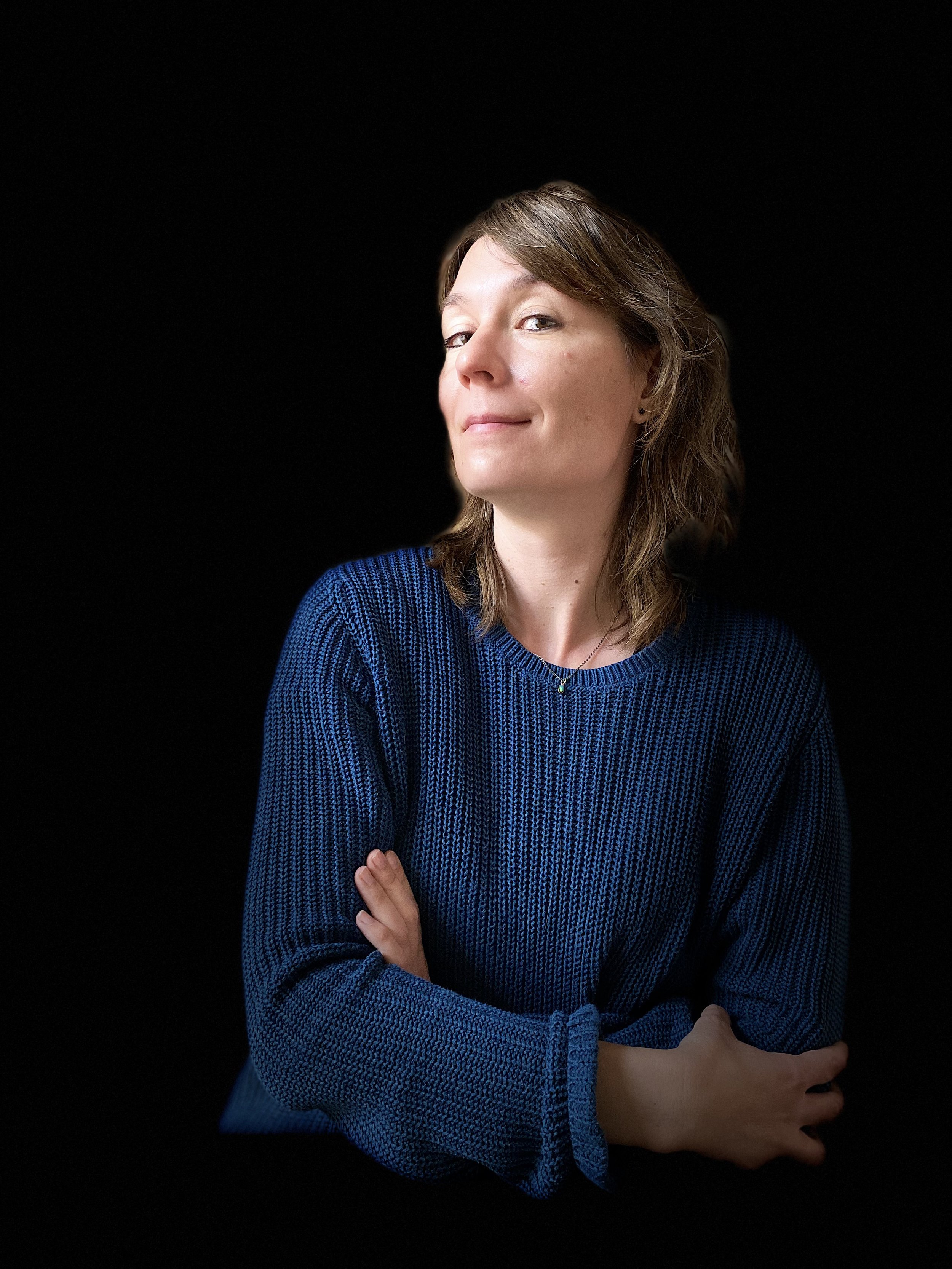 Porträt einer Frau mit braunen Haaren, die eine schwarze Hintergrund hat, trägt eine blaue Strickjacke und blickt selbstbewusst in die Kamera.