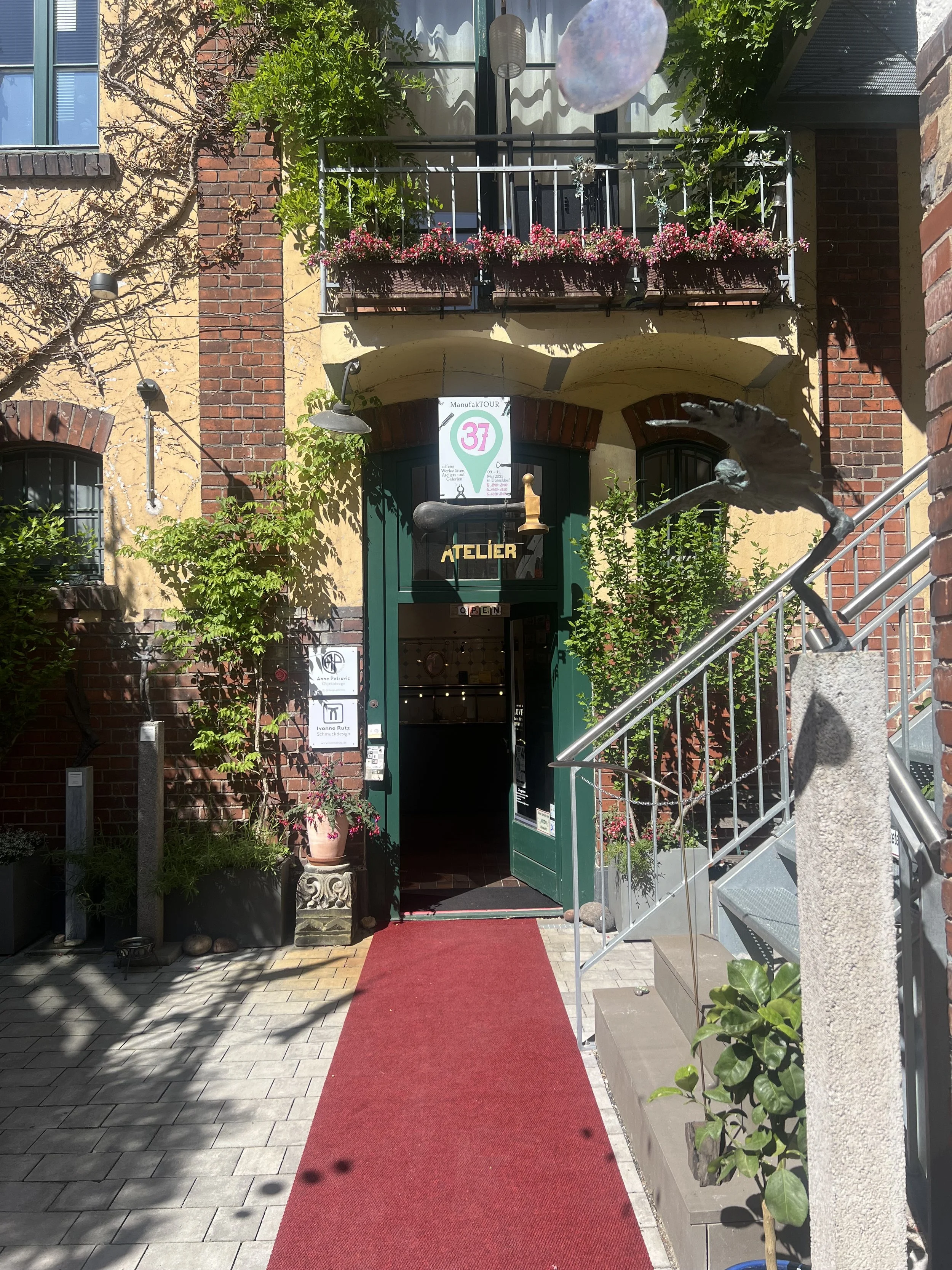 Der Eingang eines Cafés oder Ateliers mit einer roten Trittmatte, grüner Tür, Balkon mit Blumen, Pflanzen und einer Skulptur eines fliegenden Vogels