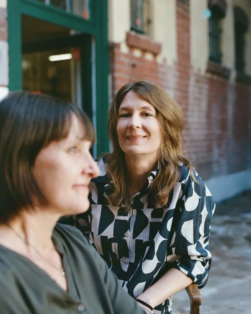 Zwei Frauen sitzen draußen vor einem roten Backsteingebäude, eine lächelt direkt in die Kamera, die andere schaut zur Seite.