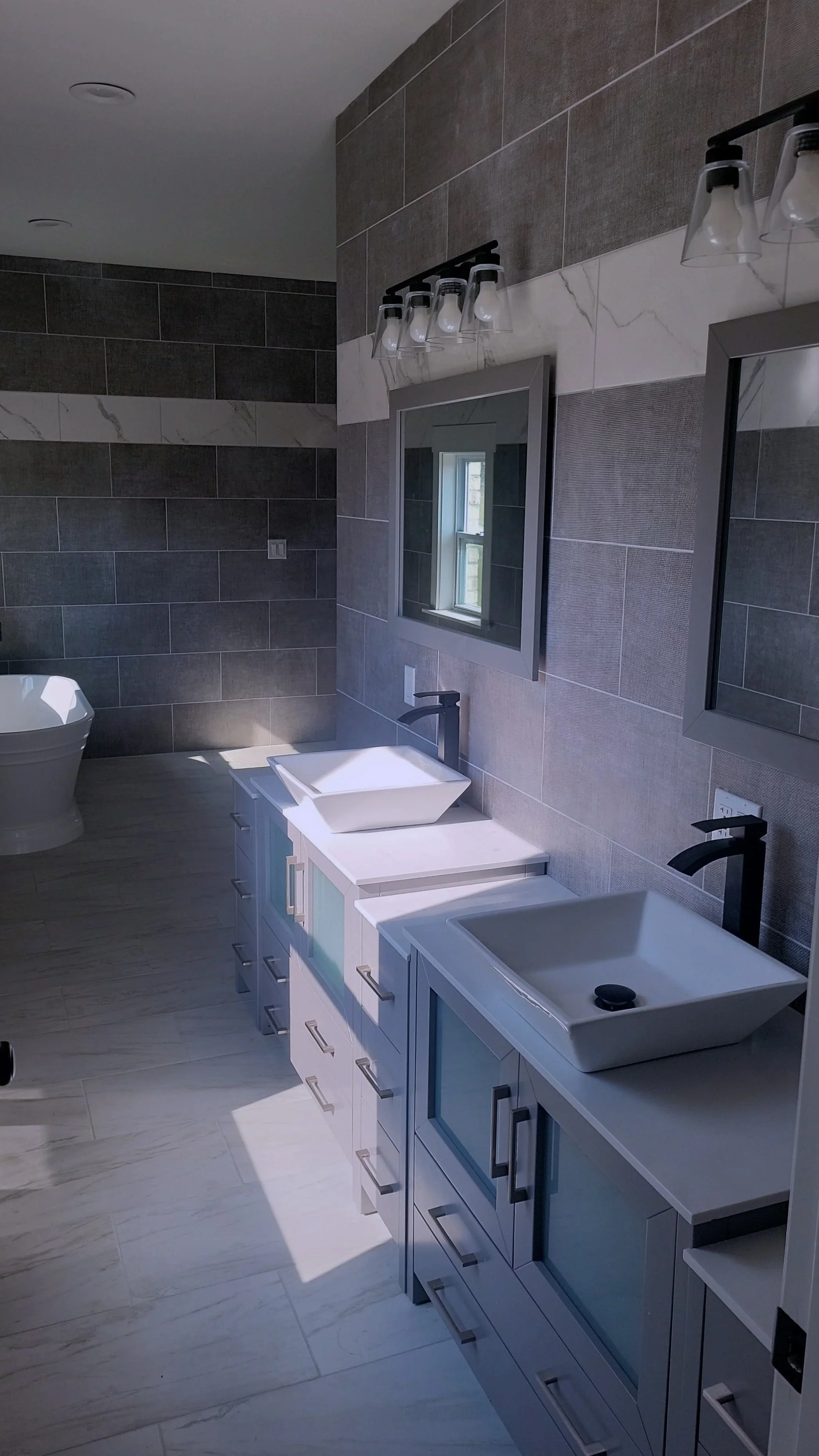 A modern bathroom with two vessel sinks on a white vanity, two mirrors above the sinks, and a bathtub in the background, with gray and white tiled walls.